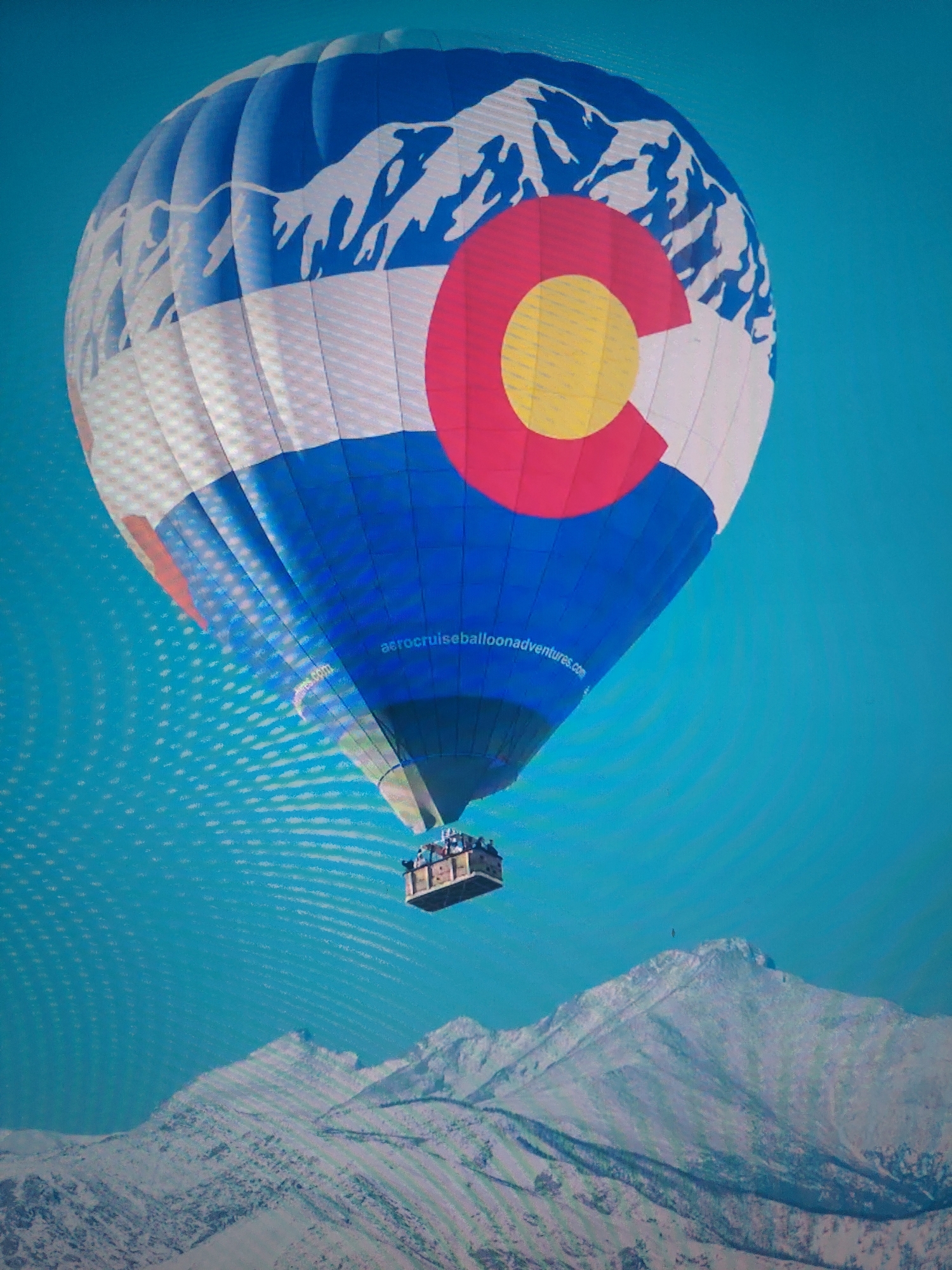 A hot air balloon, patterned with white mountains, white and red horizontal stripes, and a red "C" with a yellow circle in the center, floats in a blue sky above snow-covered mountains. A wicker basket hangs below the balloon, appearing to contain multiple people. The balloon is printed with "aerocruiseballoonadventures.com". The location appears to be a mountainous region, possibly in Colorado.