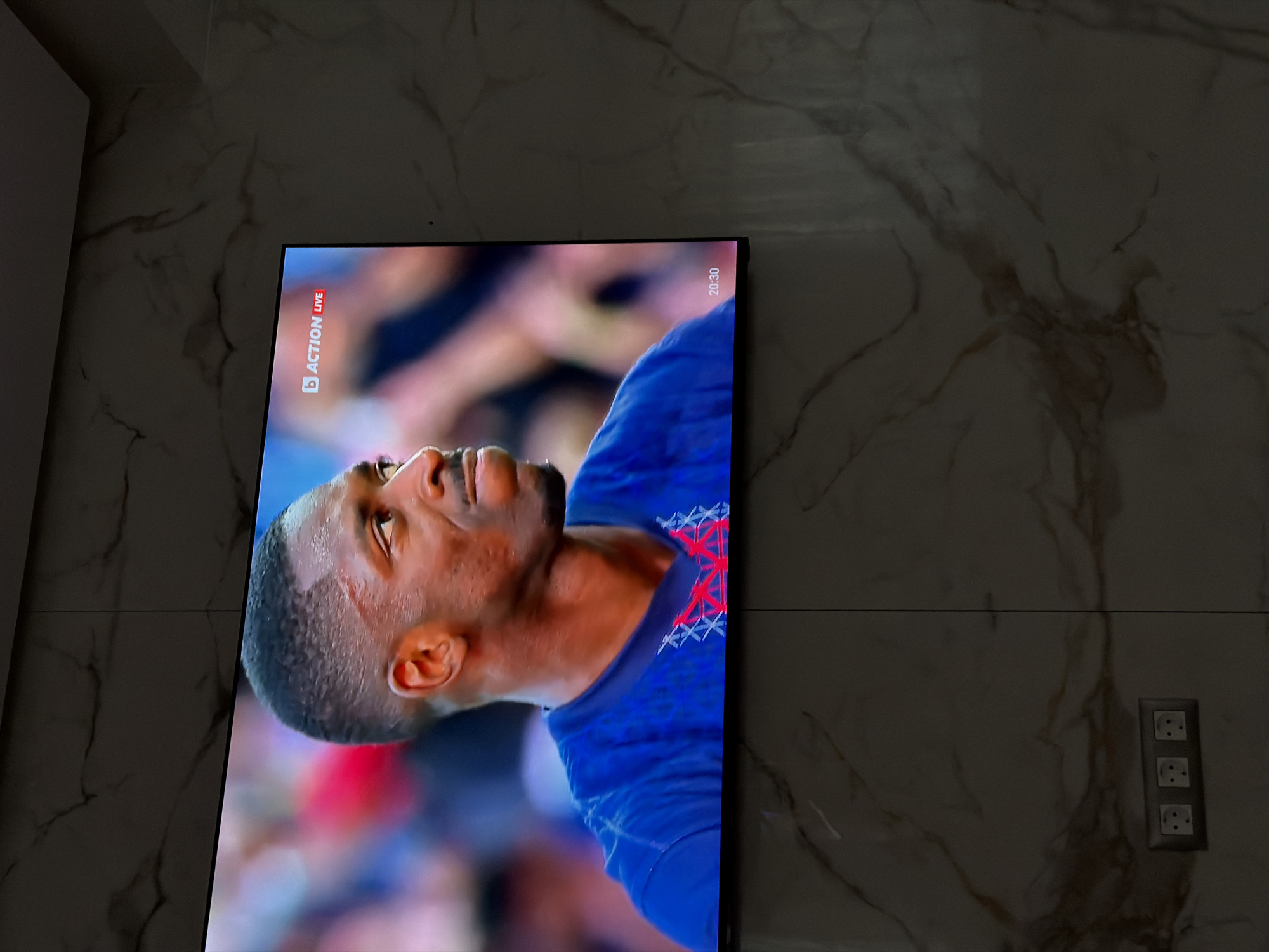 A TV screen displays a close-up of a man with dark skin, wearing a blue shirt, on a marble wall. The screen shows a live action sports event with the "ACTION LIVE" logo in the top left corner, and the time "20:30" in the top right corner. A triple power outlet is visible to the right of the TV. There is no information available to confirm if the image was taken in the city of Asenovgrad, Bulgaria.