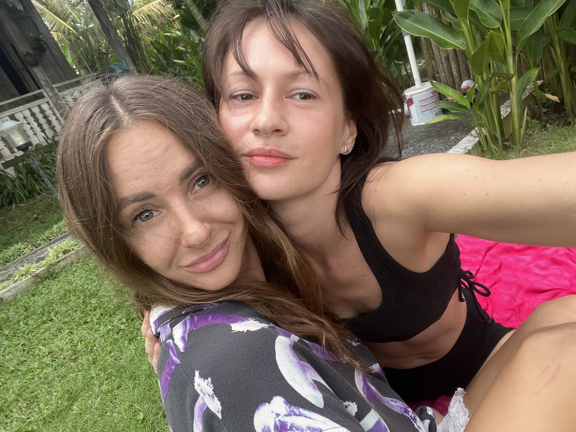 Two women pose closely for a selfie outdoors. One woman, wearing a black sports bra and shorts, is seen from the chest up. The second woman, wearing a grey and purple floral patterned top, is seen from the chest up as well. They are positioned on a bright pink blanket. Behind them is a green lawn and lush foliage, including palm trees and dense shrubbery. A building with a white fence is visible in the background. The setting appears to be Mengwi, Indonesia, based on the typical Indonesian tropical landscape.