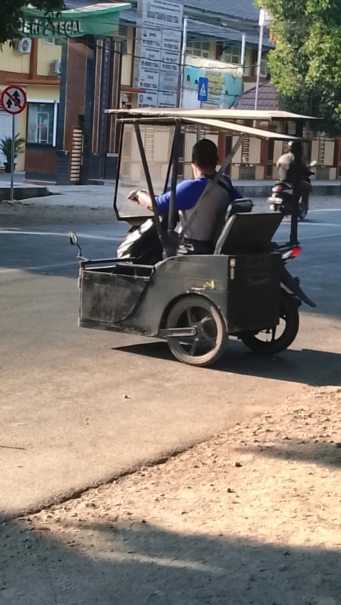 The image depicts a three-wheeled motorized vehicle, commonly known as a "becak motor" or "ojek becak," with a driver and a passenger. The vehicle is black and has a covered passenger area. The driver is wearing a blue shirt and the passenger is wearing a dark shirt and dark pants. The vehicle is on a paved road, likely in a city setting, given the presence of buildings and a street sign in the background. One building in the background appears to be a government office or school with signage that includes "TEGAL" and "UNIVERSITAS". The scene appears to be an outdoor setting with trees visible in the background. The time of day appears to be daytime, with sunlight casting shadows on the road. There is another person on a motorcycle in the background. The vehicle is positioned in the foreground of the image, with the driver's back to the viewer.