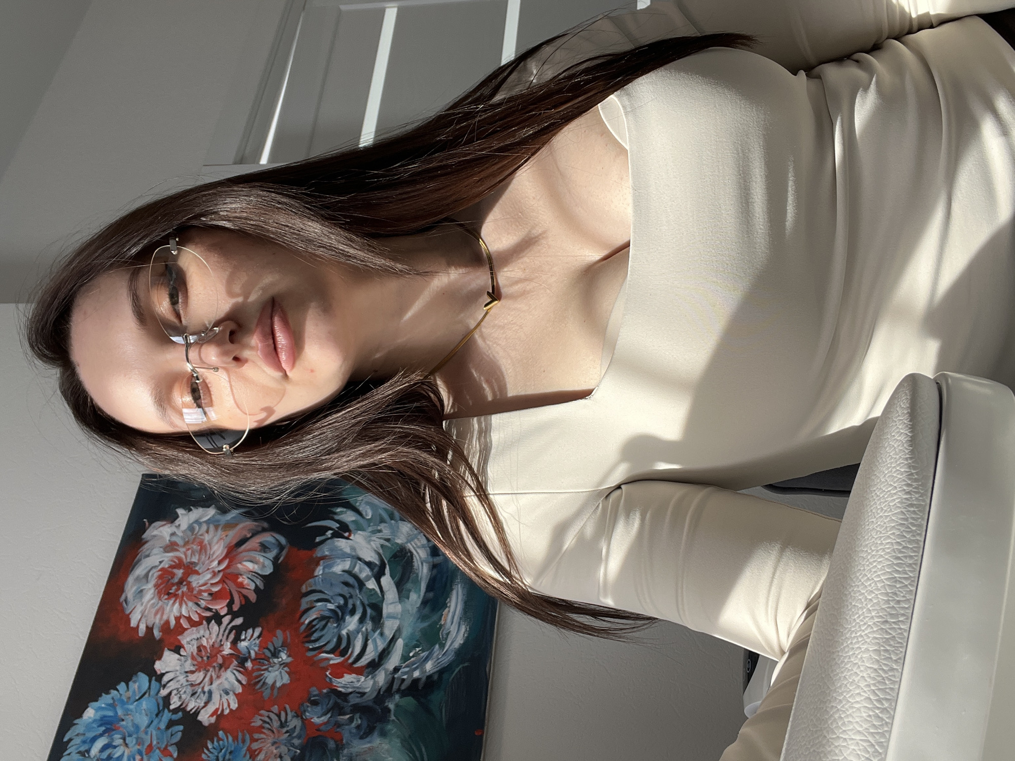 A woman is facing the camera while wearing glasses and a white long-sleeve dress. She has long, dark hair. A gold necklace adorns her neck. A painting with flowers is visible behind her, and there is a glimpse of a white door in the background. The location is possibly in Tyumen, Russia, as requested.