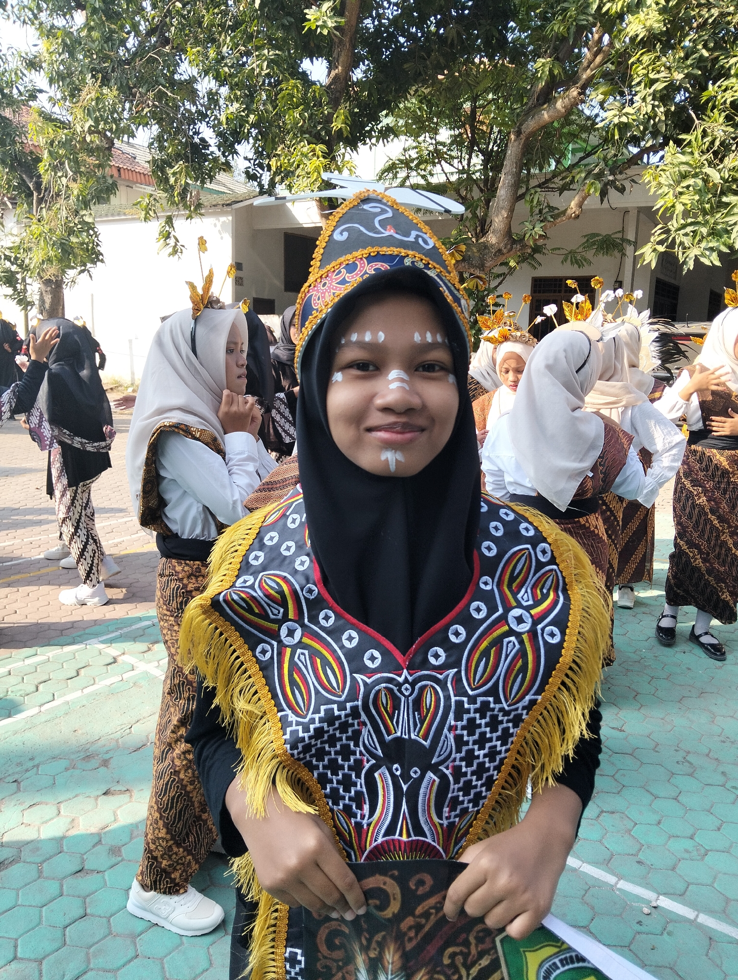 In Kranji, Indonesia, a group of young women, possibly students, are gathered outdoors on a paved area, likely part of a school campus. The foreground is dominated by a young woman wearing a black hijab and a black long-sleeved shirt. She has white face paint on her cheeks and forehead, resembling traditional markings, and is smiling at the camera. She is holding a patterned dark object, possibly a folder or program, in her hands.
She is adorned with an elaborate, colorful vest or sash over her shirt, featuring intricate patterns in black, white, red, and yellow, with gold fringe along the edges. Her lower body is partially visible, showing a black sash and a patterned brown and gold batik skirt or sarong. She is wearing white sneakers.
Behind her, other young women are also dressed in traditional attire. Many wear light-colored hijabs, some with decorative gold headpieces featuring leaf-like or feather-like elements. They are also wearing batik skirts or sarongs with similar brown and gold patterns. One woman to her left wears a black hijab and a black top with a batik sarong. The background reveals a building with white walls and a tiled roof, and lush green trees with dappled sunlight filtering through their leaves. The ground is paved with hexagonal tiles, some of which are painted green, forming a grid. The overall scene suggests a cultural event or performance taking place outdoors.