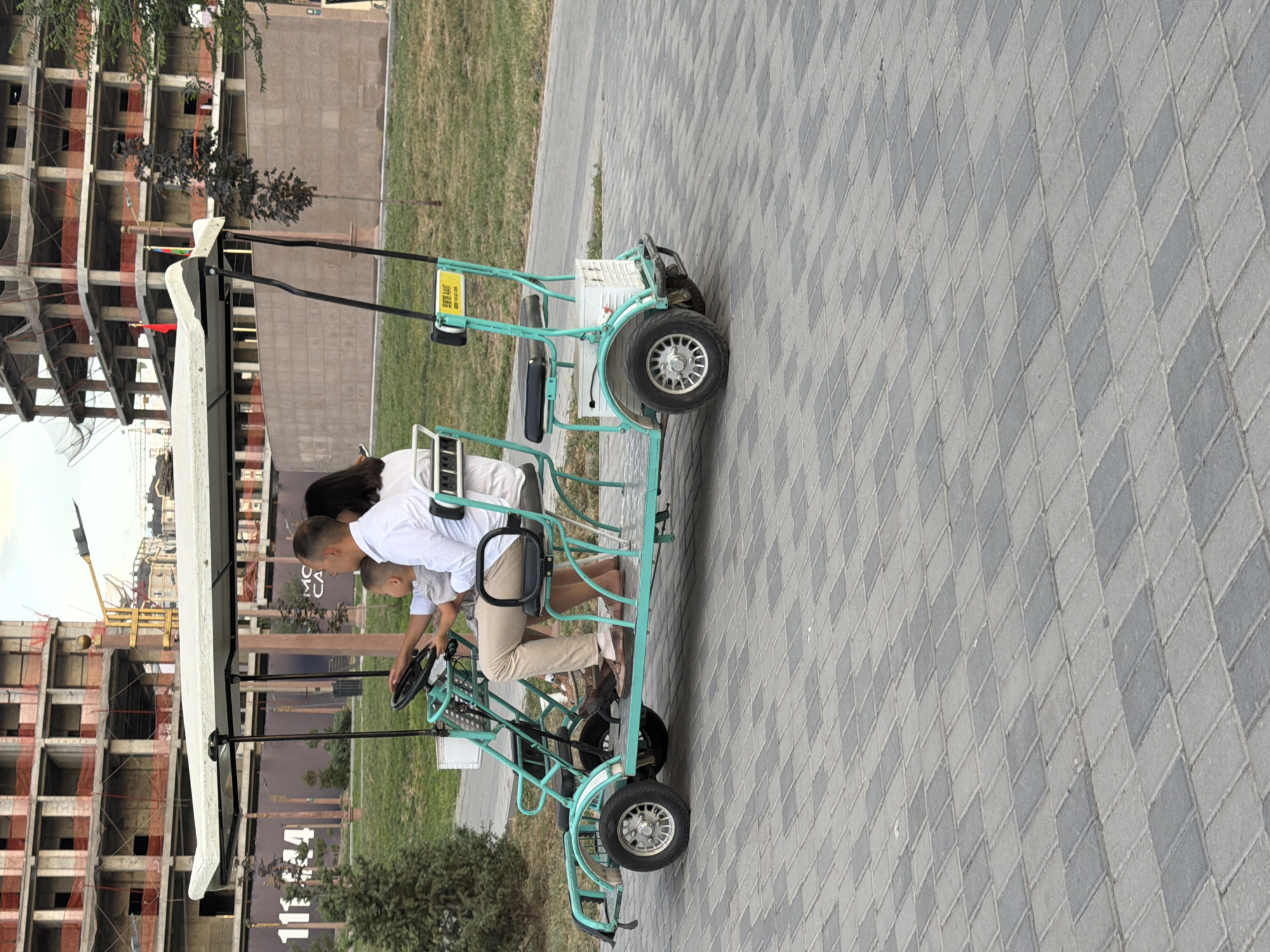 An outdoor, eye-level, medium shot captures a family of three riding in a turquoise, four-wheeled golf cart along a patterned gray and dark gray brick pathway. The cart has a white canopy top and black seats. A man in a white long-sleeved shirt and tan pants is in the driver's seat, holding the steering wheel. A young boy in a gray t-shirt sits in front of him, and a woman in a white top is seated behind the man, facing away from the camera. Her dark hair is visible. A white crate is attached to the rear of the cart.
In the background, a large, unfinished building with exposed concrete floors and red netting on its exterior is under construction. A street light and several trees are also visible. The ground is covered with green grass and a gray asphalt path to the right of the brick walkway. The sky is overcast.