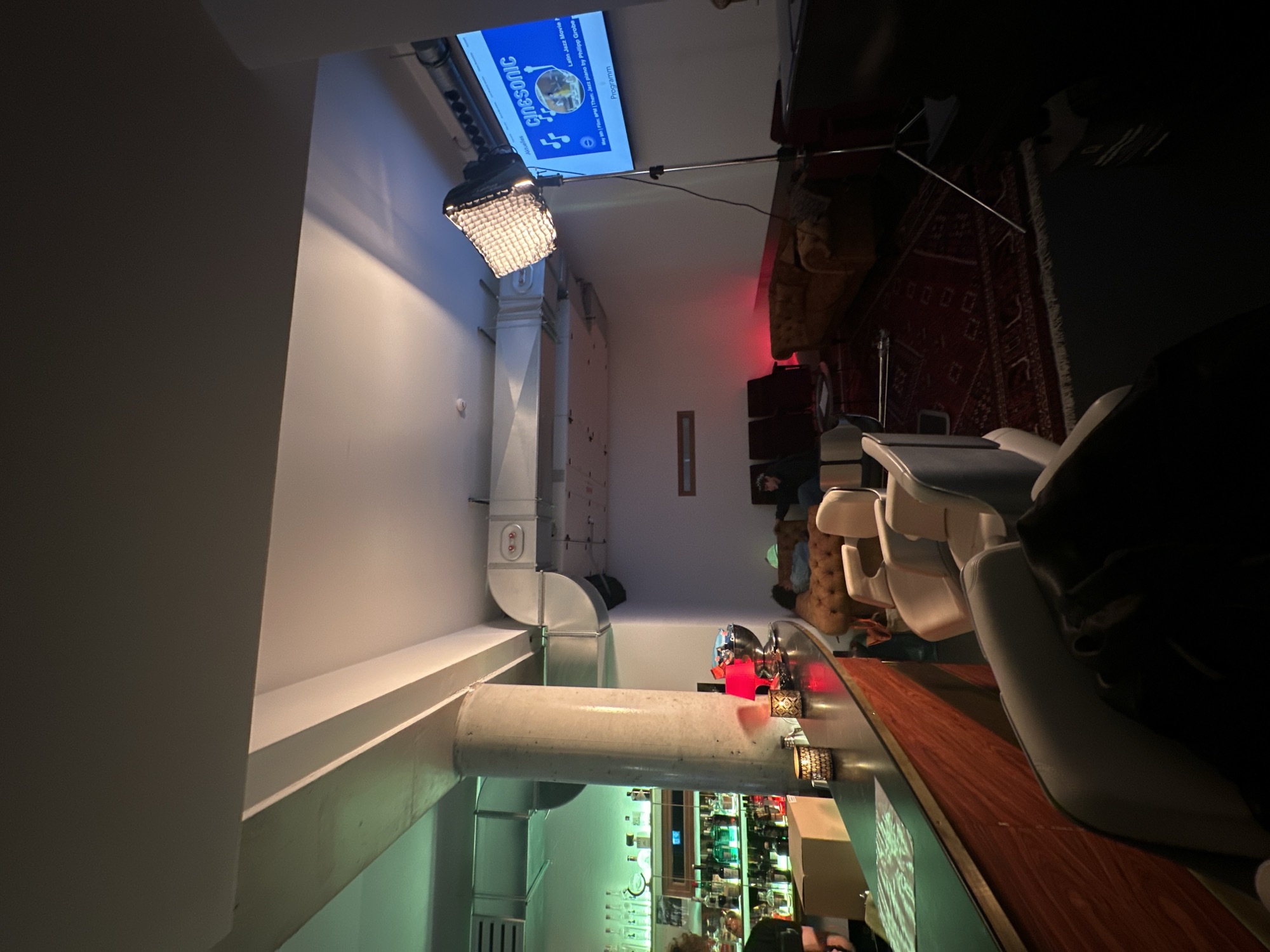 The image captures an indoor bar or lounge setting, likely in the evening or at night, characterized by dim, atmospheric lighting. The venue appears to be hosting an event.
Prominently featured on the ceiling is a white projector screen displaying event information. Visible text on the screen includes "Aktuelles" (Current/News), "CINESONIC," "Latin Jazz Movie," and details a program for "May th | Film: BPM | Then: Jazz piano by Philipp Grobe," followed by "Programm" (Program). A large light fixture with a grid diffuser, attached to a stand, projects light towards the screen. Exposed white and silver air ducts run along the ceiling and walls, contributing an industrial aesthetic to the space.
To the right, a wooden bar counter is visible with several bar stools or chairs. Behind the counter, shelves filled with bottles create a typical bar backdrop. Further back, a lounge area features a patterned red rug and upholstered seating, where several indistinct individuals are seated or standing, appearing to socialize or observe the event.
The lighting is predominantly artificial, with the bright projector screen contrasting with the dim surroundings. A noticeable red light emanates from the background, adding to the ambiance. Small decorative lights or candles are placed on the bar counter. The overall impression is that of a cozy yet modern venue hosting a cultural event, potentially in Berlin, Germany.