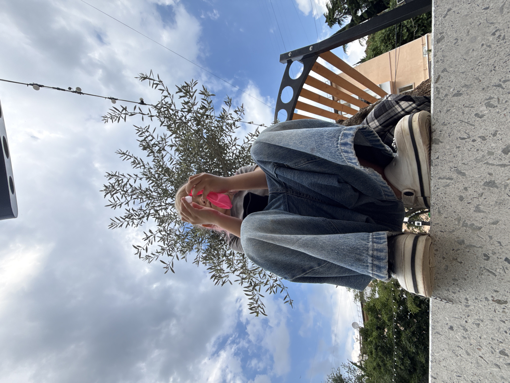 A low-angle, wide shot shows a person sitting on a concrete ledge in an outdoor setting. The person is wearing a grey t-shirt, very baggy blue jeans, and white chunky platform sneakers. They are holding a bright pink face mask up to their face, obscuring their eyes and nose. Behind the person is a small tree with sparse branches and foliage. Above the tree, string lights with small white bulbs are visible against a cloudy blue sky. To the right of the person, a wooden bench with circular cutouts is partially visible. In the background, trees and buildings are visible. The overall impression is one of a casual, candid moment captured outdoors, with the person's obscured face and the wide, slightly distorted perspective creating a unique and engaging composition.