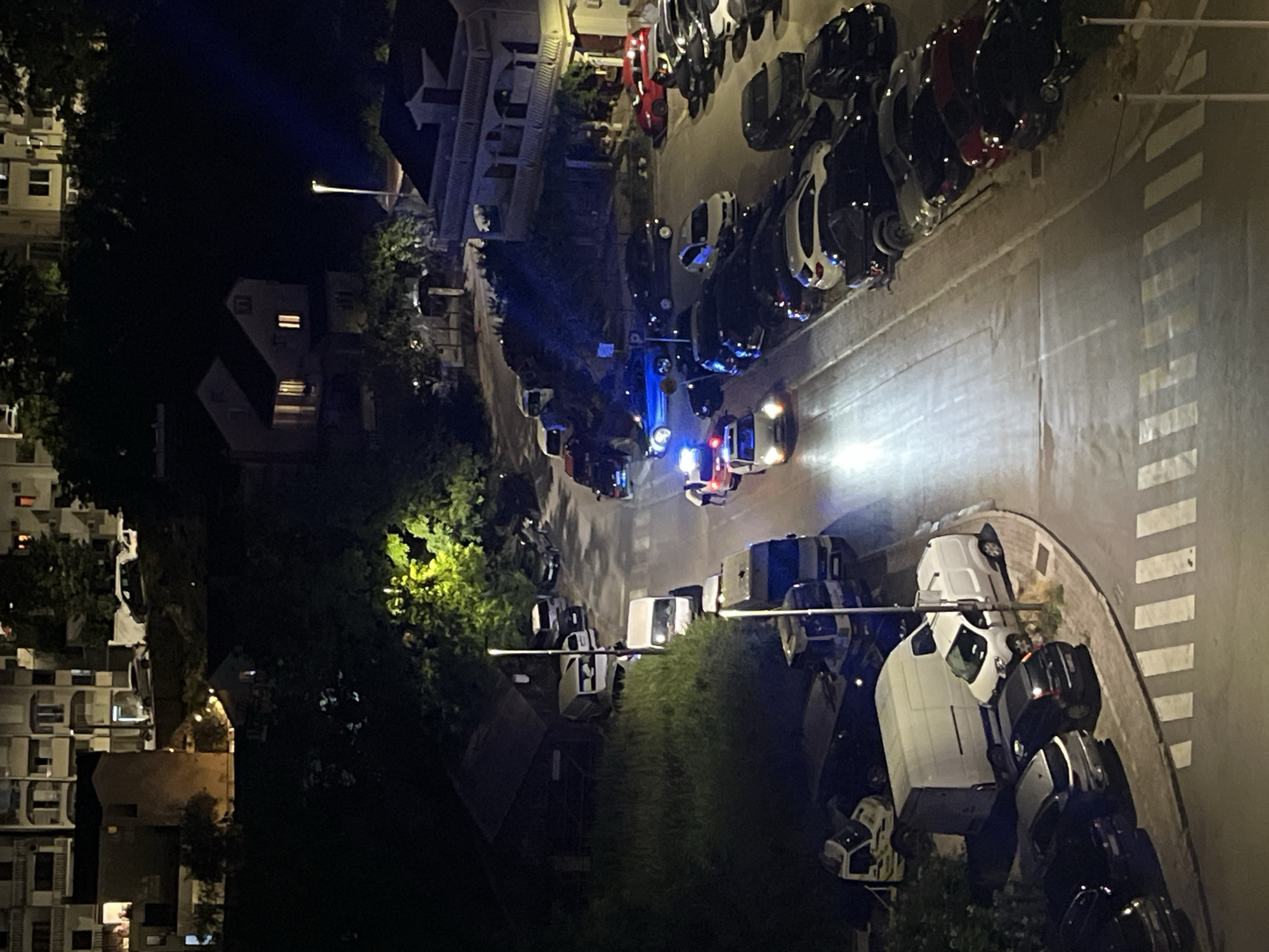 An aerial nighttime view captures a residential street in Budva, Montenegro, with multiple vehicles present, including police cars. Several police cars with flashing blue lights are stopped in the middle of the road, and two individuals in uniform are visible near one of the vehicles. The street is lined with parked cars on both sides, and a white van is parked to the left. The surrounding environment features residential buildings with illuminated windows, trees, and streetlights.
