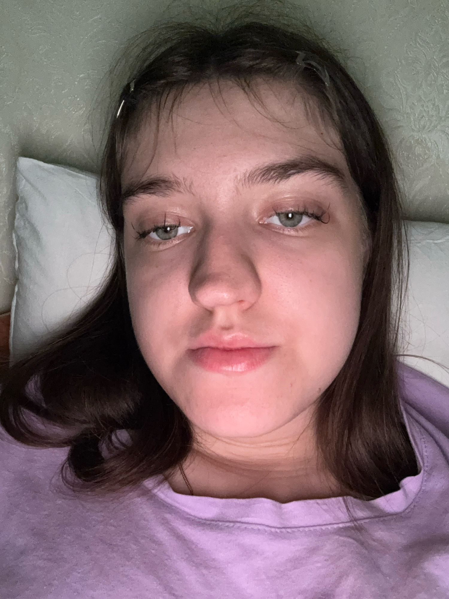 A young female is lying on a pillow, looking upwards towards the camera. She has dark brown hair, light skin, and green eyes. She is wearing a light purple t-shirt. The background appears to be a wall with a textured pattern. The image is a close-up shot, focusing on her face and upper torso. There is no indication of the specific location within Zavolzhye, Russia, or any activities or interactions occurring.