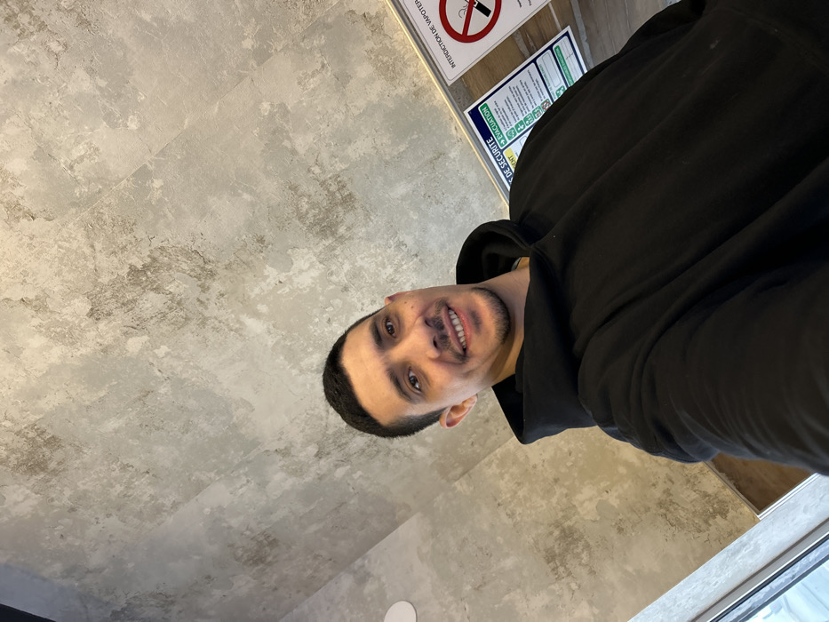 A male individual with short dark hair, light stubble, and a smiling expression is depicted in a selfie, wearing a black hooded sweatshirt. The subject is positioned indoors against a mottled gray and brown textured wall, which appears to be concrete or a similar industrial finish. To the left, a vertical window or glass panel is partially visible, showing a blurred exterior. On the right side of the frame, two signs are affixed to the wall above a wooden or wood-patterned paneling. One sign is blue and white, displaying text including "DE SECURITE" (safety) and "EVACUATION." Adjacent to it is a circular sign with a red border and diagonal line, illustrating a vaping device with the text "INTERDICTION DE VAPOTER" (Prohibition of vaping). The French language on the signage confirms the location within Schiltigheim, France.