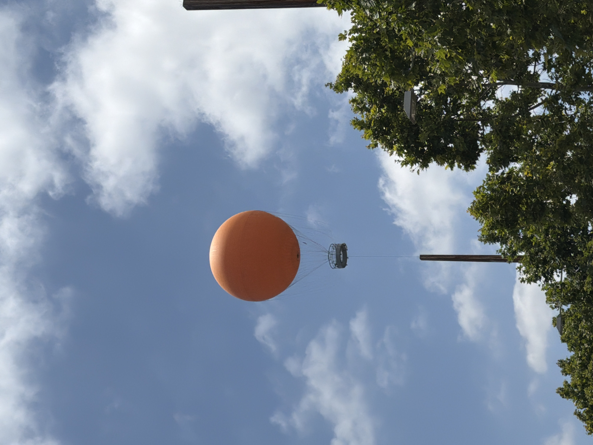 An orange hot air balloon with a circular basket is suspended in a blue sky with scattered white clouds. The balloon is tethered to a dark brown pole that extends from the bottom of the frame. Lush green trees with visible branches and leaves occupy the lower right portion of the image, partially obscuring another dark brown pole and a street light fixture. The scene appears to be in Irvine, United States of America.