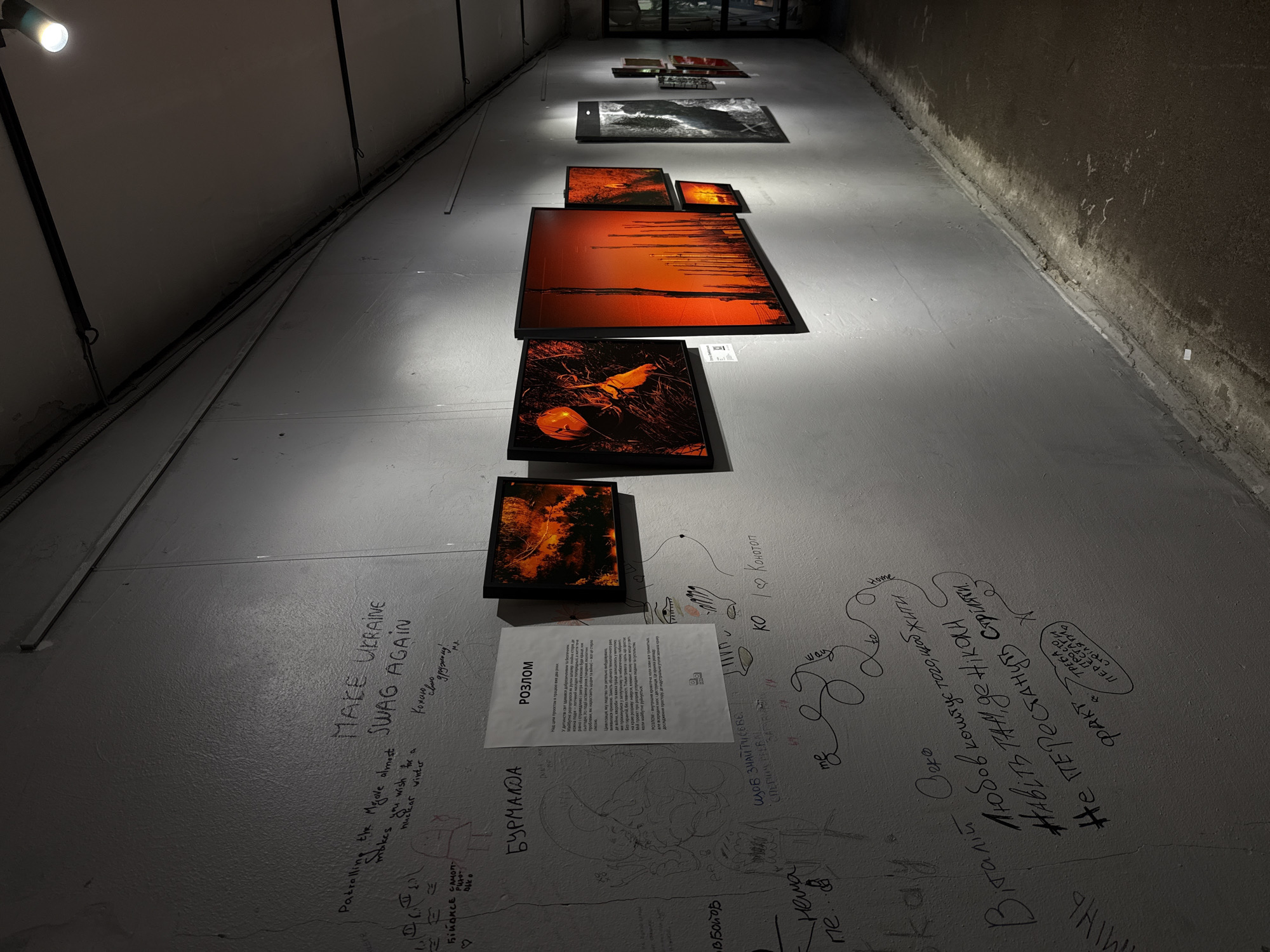 The image shows an interior view of a dimly lit corridor, likely part of an art exhibition space. The walls are painted a rough, textured gray and are covered in graffiti and posted flyers. In the foreground on the left, there is handwriting and some sketched figures. A white flyer with Ukrainian text is prominently displayed on the wall. Further down the wall, a series of framed photographs are hung, predominantly featuring warm, orange and red tones, suggesting images of landscapes or abstract art. The floor is concrete and appears somewhat worn and damp in places. The overall impression is of an underground or unconventional artistic venue. Without further information or context, it is not possible to definitively confirm the location as Kyiv, Ukraine, though the presence of Ukrainian text on the flyer and in the graffiti could suggest this.