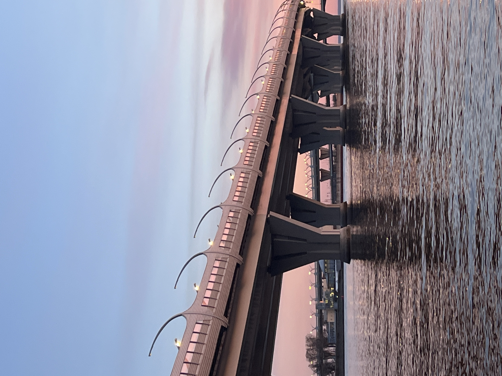 The image captures a modern bridge spanning a body of water at twilight, likely in Saint Petersburg, Russia. The bridge, extending diagonally across the frame from the left foreground to the right background, features a prominent elevated roadway supported by robust concrete pillars. Along its length, numerous distinctive, curved lighting fixtures are installed, casting a warm glow as they are illuminated.
The sky above the bridge displays a soft, gradient blend of pastel colors, transitioning from light blue on the left to delicate pinks and purples towards the right, indicative of either dawn or dusk. The calm water beneath the bridge reflects these gentle sky hues and the bright lights from the bridge, creating shimmering, vertical streaks on the surface. Distant elements of a shoreline or cityscape, adorned with faint lights, are visible through the arches of the bridge's supports. The scene conveys a sense of peacefulness and serenity, showcasing the architectural beauty of the bridge against the colorful twilight backdrop. No visible text is present in the image. The bridge's design is characteristic of the Western High-Speed Diameter (WHSD) in Saint Petersburg.