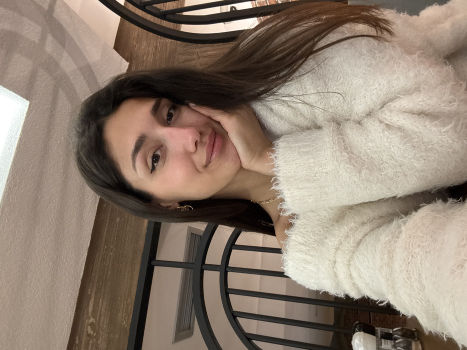 An image taken in Zankovići, Montenegro, depicts a young adult female in a close-up, chest-up shot. The subject has long, dark brown hair and is wearing a white, off-shoulder, fluffy knit sweater. She has light makeup, including black winged eyeliner and a subtle lip gloss. Gold-colored hoop earrings are visible on both ears, and a thin gold-colored chain necklace is around her neck. Her right hand is positioned with fingers resting against her cheek. She is looking directly at the camera with a slight smile. The background suggests an indoor setting, likely a restaurant or cafe, featuring a section of a light-colored ceiling with a square recessed light fixture. Directly behind the subject's left shoulder, part of a dark, possibly metallic, vertical lattice or decorative screen is visible. To the bottom left, partially visible on a surface, are what appear to be two small, clear glass shakers with white tops, commonly used for salt and pepper. Further back on the left, a light-colored wall with a horizontal vent is visible above a dark horizontal beam. On the right side of the frame, another segment of the dark metallic lattice/screen is present.