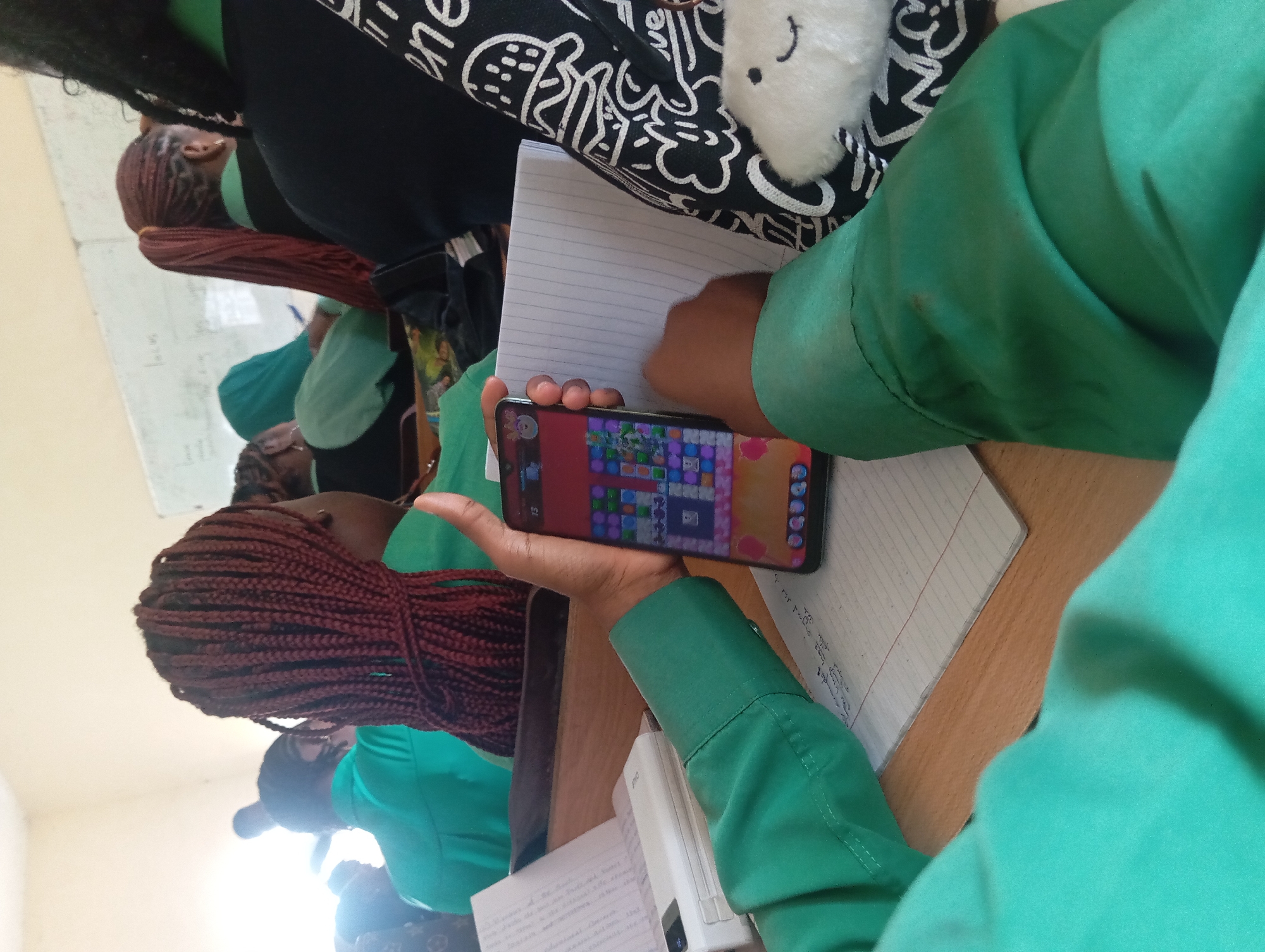 In an indoor classroom setting, a student in a green uniform is holding up a smartphone, playing a game. The phone displays a colorful game interface with matching tiles. The student's left hand is visible, gripping the bottom of the phone, while their right hand supports it from the side. The student's arm is covered by the sleeve of their green uniform. In the background, other students are seated at desks, some with their backs to the camera, wearing similar green uniforms. The environment appears to be a school, with desks and a whiteboard visible in the distance. A notebook and a white electronic device are on the desk in front of the student. A black bag with a white graphic design and a fuzzy white keychain is partially visible to the right of the phone. The lighting suggests an overcast day or an indoor space with diffused light.