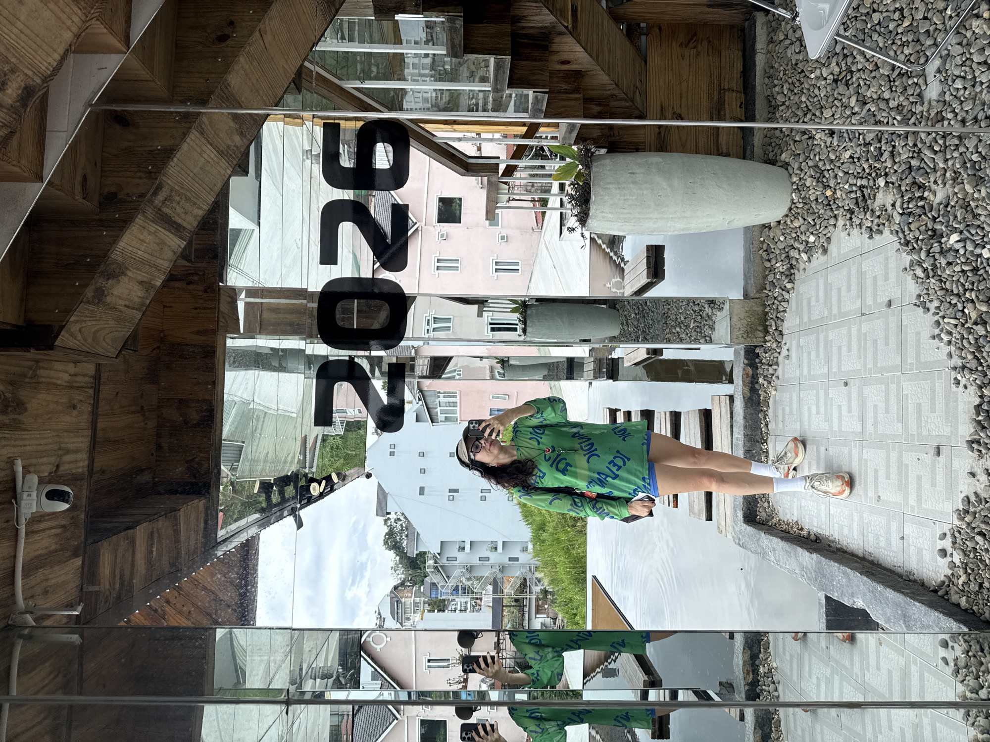 A young Asian woman stands in front of a mirrored wall, taking a selfie. She is wearing a long-sleeved green sheer top with blue lettering, blue shorts, white socks, and beige and orange sneakers. She has on a visor and headphones. The mirrored wall reflects a city scene with buildings and lush greenery, suggesting the location is Da Lat, Vietnam, a city known for its scenic hills and French colonial architecture. The woman's reflection is also visible in the water feature in front of her, with wooden planks forming a walkway across it. Behind her, a wooden staircase and potted plants are visible. The number "2025" is prominently displayed on the mirrored wall.