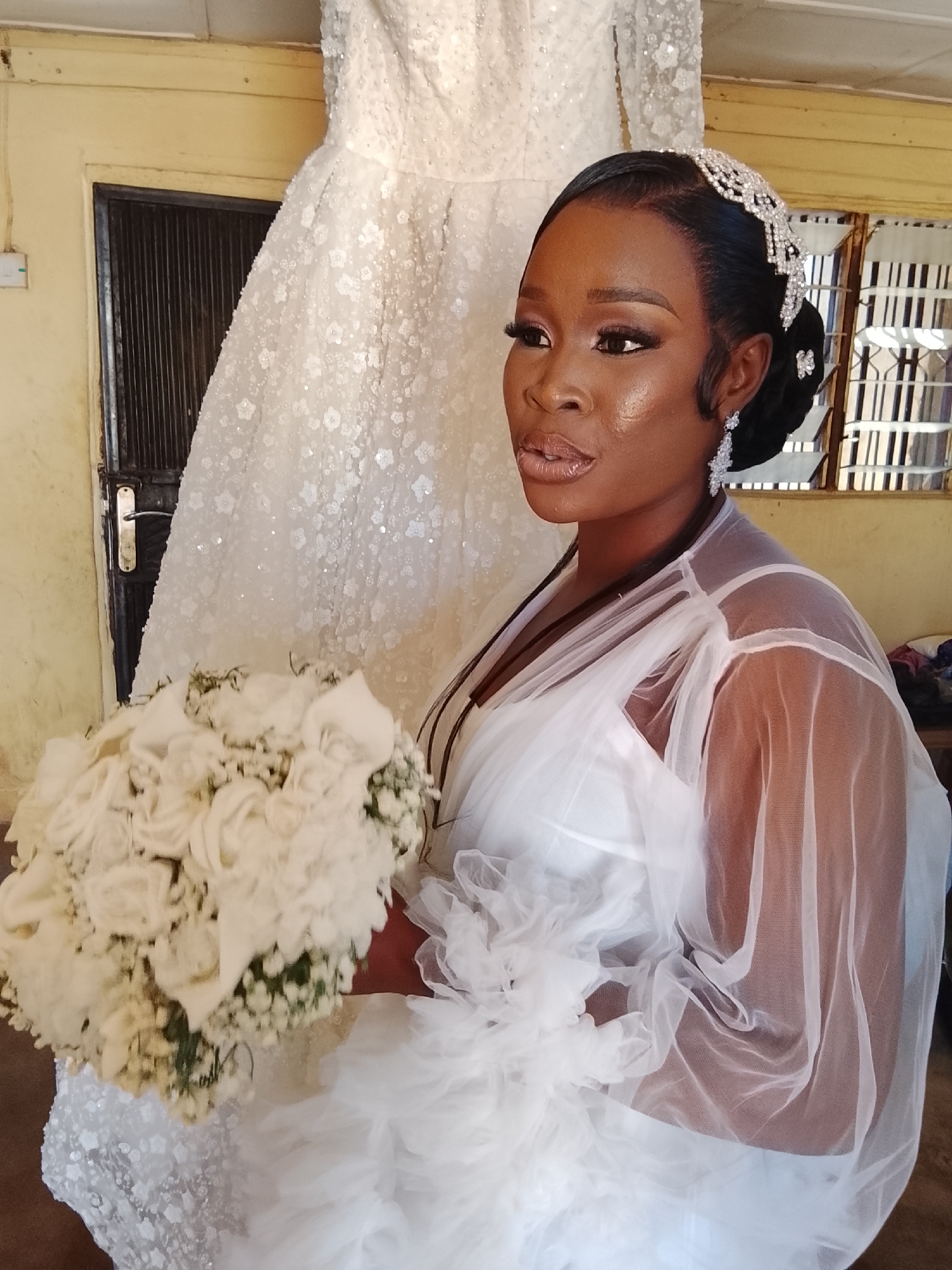 A medium shot captures a dark-skinned woman in a white dress, holding a bouquet of white flowers. Her hair is styled back and adorned with a jeweled headband. She is wearing dangling earrings. A white wedding dress hangs in the background next to a dark metal door. The location is in a room with mustard colored walls, in the city of Unknown, Unknown.