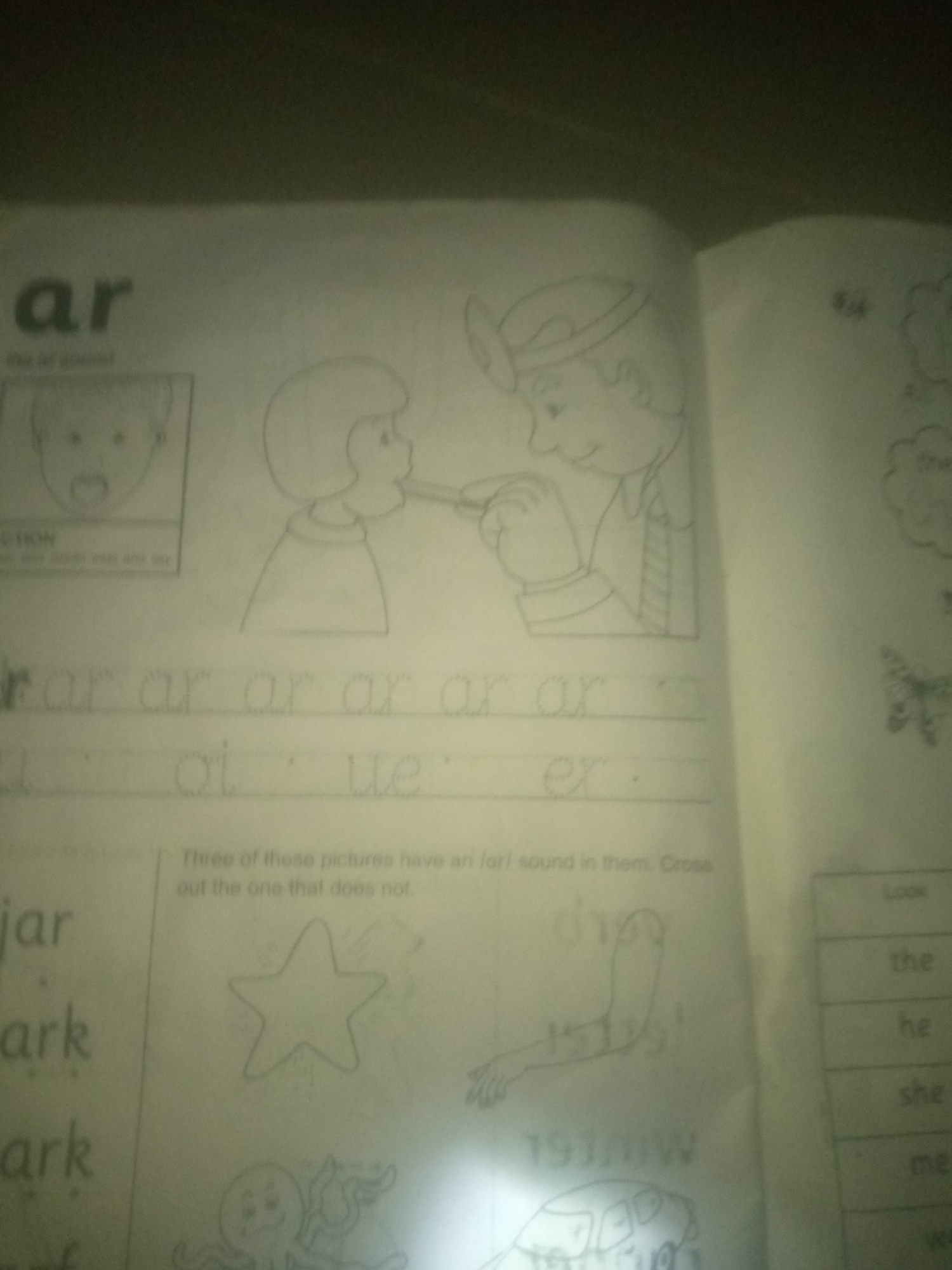 The image is of a page from a children's workbook, likely for early literacy education. The page focuses on the "ar" sound in words. In the upper section, there are two cartoon drawings. The left drawing shows a baby's face with an open mouth, illustrating the "ar" sound. The right drawing depicts a doctor examining a child's throat with a tongue depressor. Below these drawings, the letters "ar" are written multiple times in a large, dotted font, suggesting a tracing activity. Further down, other letter combinations like "oi" and "ue" are also present in a similar dotted style. Below this, a sentence reads, "Three of these pictures have an /ar/ sound in them. Cross out the one that does not." To the left, words like "jar," "ark," and "ark" are written, again in dotted font, implying tracing or writing practice. On the right side of the page, there are small drawings of a butterfly, a star, a human arm, and an octopus, and a car which might be related to the exercise. A small table on the far right contains words like "Look," "the," "he," "she," and "me." The overall environment appears to be indoors, with the lighting suggesting artificial light sources, potentially from a lamp or flash, casting a slight glare on the page. There is no visible information or context within the image that would suggest a specific location within Jauro Yinu, Nigeria.
