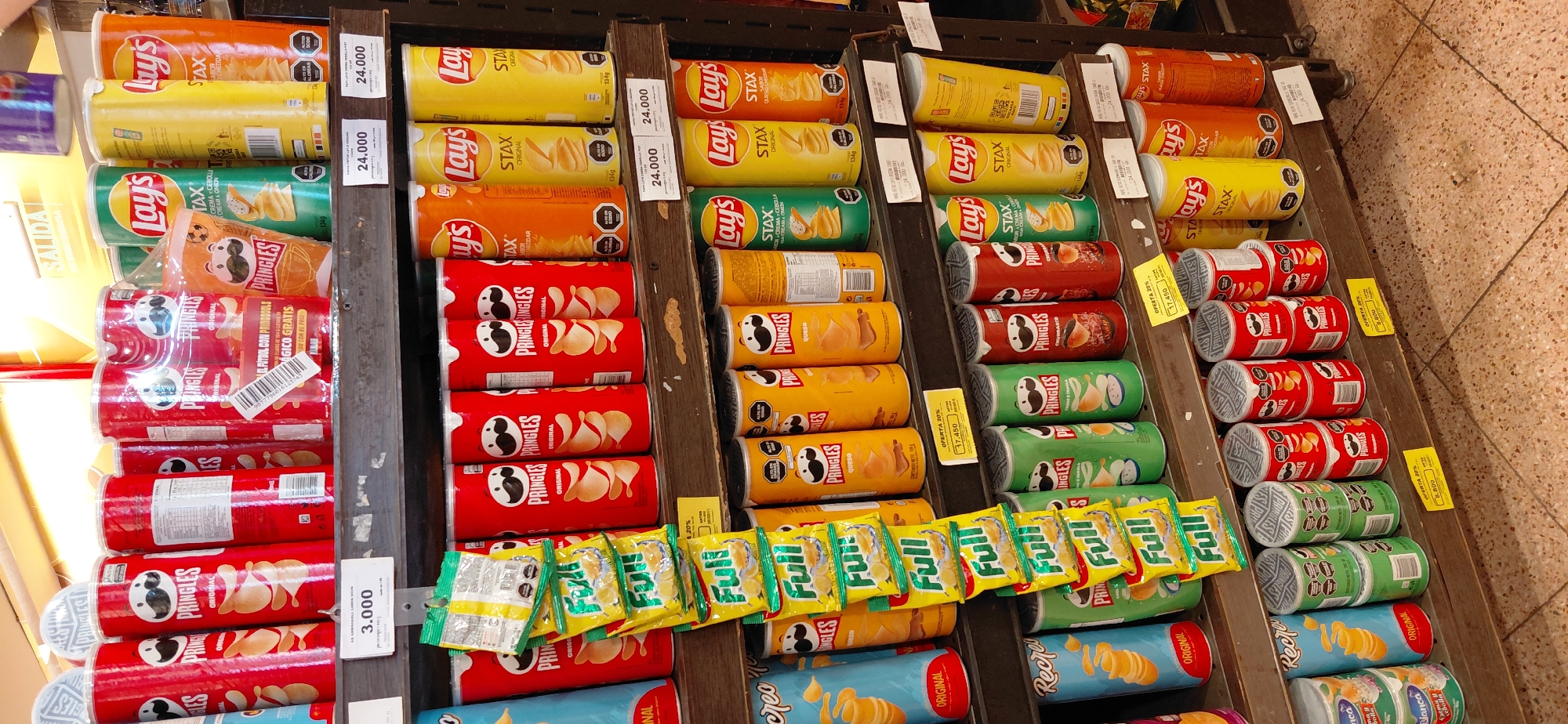 The image shows shelves in a store stocked with various brands of potato chips, including Pringles and Lay's. The chips are packaged in cylindrical cans and bags, in a variety of flavors and colors. Some cans are stacked on top of each other. There are price tags visible on the shelves, with prices such as 3.000, 24.000, and 17.450. The lighting suggests an indoor commercial setting. The location is identified as Encarnacion, Paraguay, implying this is a retail environment within that city. There are no people visible in the image.