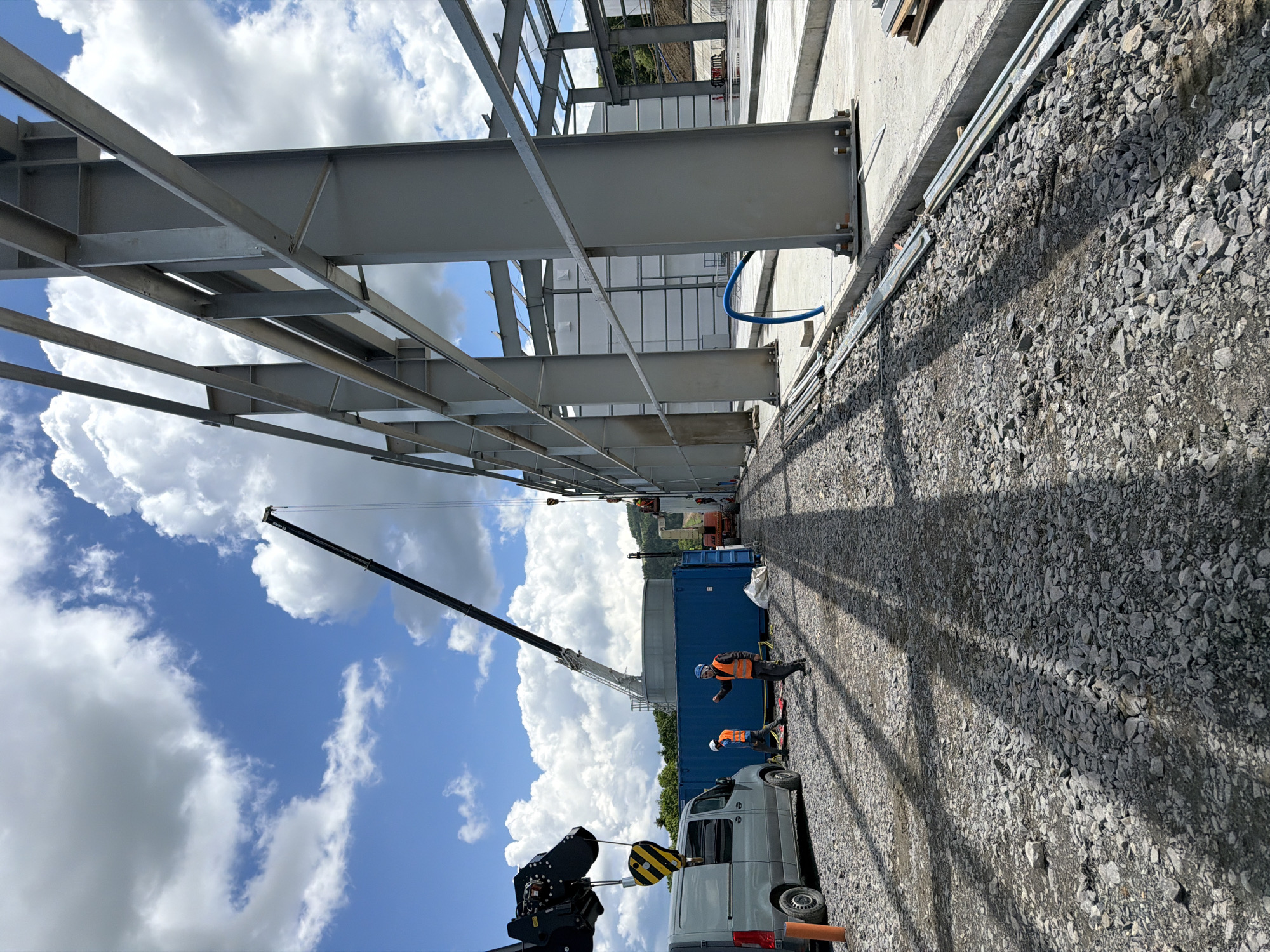 This image depicts a construction site in Stróża, Poland, focused on the erection of a large steel-framed structure. In the foreground, a gravel-covered area is visible, with shadows of the steel beams cast across it. To the left, a grey van is parked. Two construction workers, wearing high-visibility vests and hard hats, are present near the van and a large blue shipping container. One worker is gesturing while the other is walking.
A large crane is actively engaged in lifting materials, with its boom extending upwards against a backdrop of a bright blue sky dotted with white, fluffy clouds. The primary subject of the image is the developing steel framework of the building, characterized by its large grey vertical beams and horizontal supports. In the background, parts of the partially constructed building's exterior are visible, along with more heavy machinery, suggesting ongoing construction activity. The overall environment is indicative of an industrial or large-scale construction project in an outdoor setting.