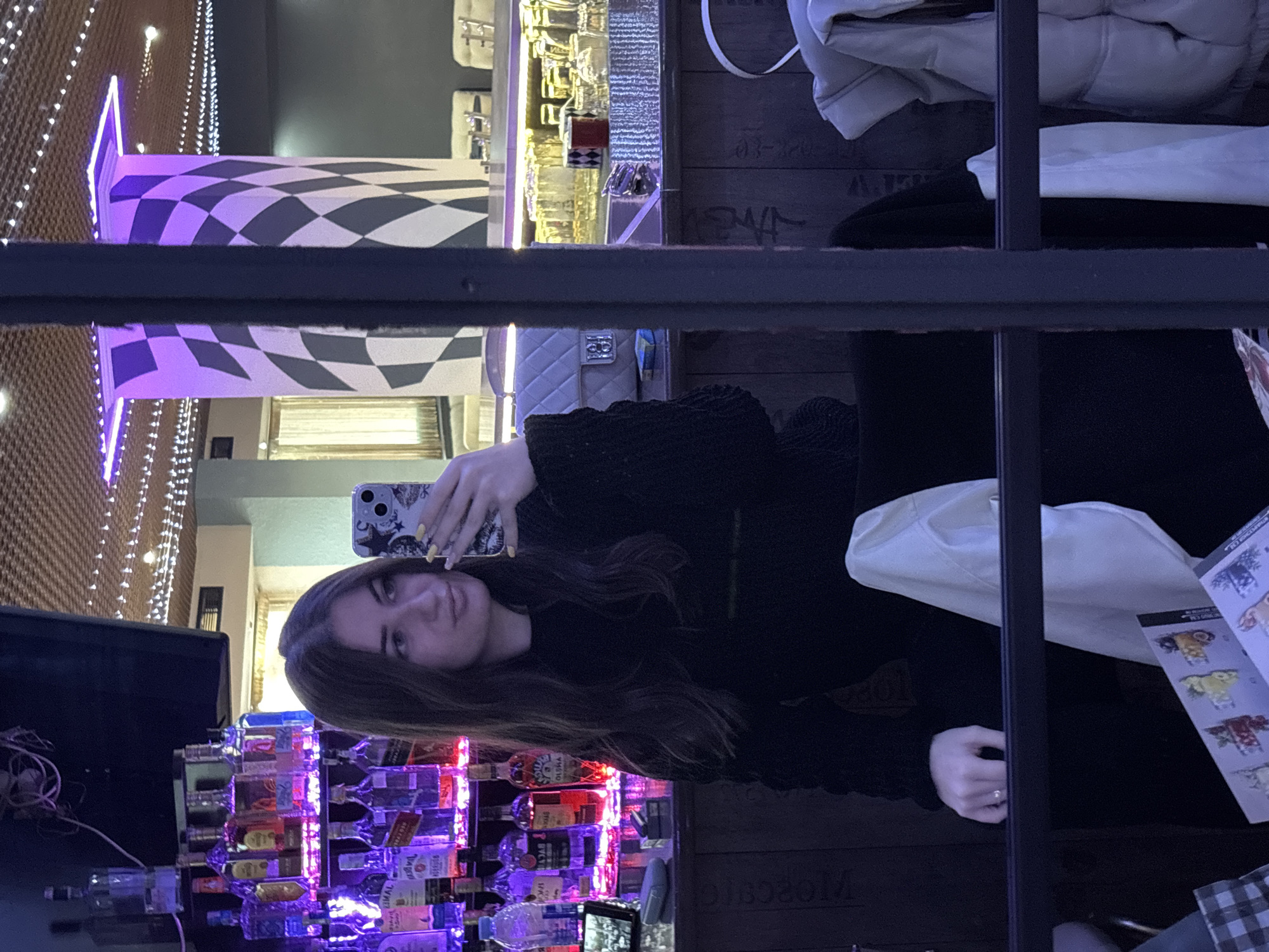 A woman is taking a selfie at a bar in Chirpan, Bulgaria. She is sitting at the bar, which is stocked with various bottles of alcohol illuminated with colored lights. A checkered design is visible on the ceiling. The woman has long, dark hair and is wearing a black sweater. A menu is visible on the table. A jacket is hanging over the back of the chair.