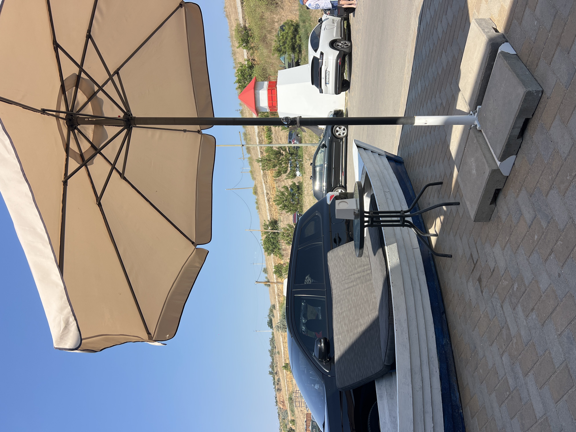 In Fontanka, Ukraine, on a sunny day, a beach umbrella shades a scene. In the foreground, a white rowboat with blue trim holds a dark grey cushion. Beside it, a small black metal table with a single item on it stands on a paver patio. Behind the boat, a black SUV is parked. Further back, a white lighthouse-shaped structure with a red roof is visible. To its right, a white SUV is parked. In the background, cars are parked on a paved lot, with a grassy, sloped hill dotted with trees and power lines behind them. A person in a blue hat and white shirt is walking away from the white SUV.