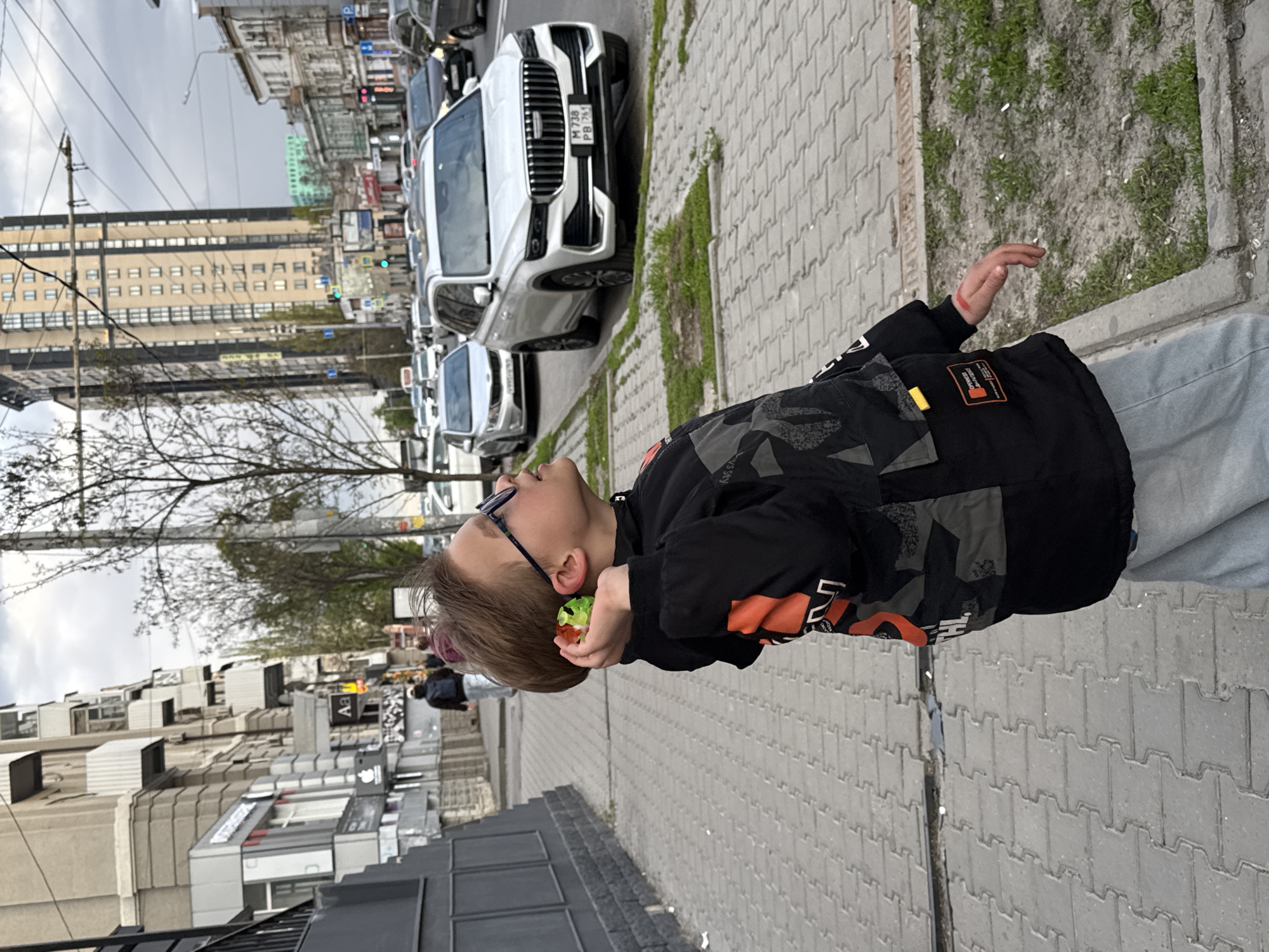 The image features a young boy in an urban setting, likely in Rostov-on-Don, Russia. The boy, with light brown hair and wearing glasses, is dressed in a black jacket with orange and camouflage-patterned accents, and light blue jeans. He is looking upwards with a slightly open mouth, holding a small, multi-colored (green, red, and yellow) spherical object near his right ear, while his left hand is raised.
He stands on a sidewalk paved with grey interlocking pavers, with a strip of dirt and sparse green growth separating it from the street. The street is lined with parked vehicles, including several white SUVs. One prominent white SUV displays a Russian license plate reading "M 738 PB 761," with "761" corresponding to the Rostov Oblast region.
In the background, various buildings populate the cityscape, including a large, beige, multi-story building with numerous windows and another building featuring a green section on its roof. Overhead utility lines and poles are visible against an overcast or dull white sky. Trees with bare branches suggest late winter or early spring. Text on the boy's jacket includes partial words such as "7" and "TH" on an orange patch, and "SS Y" on a black section. The scene captures a moment of observation or play in a typical city environment during daytime.