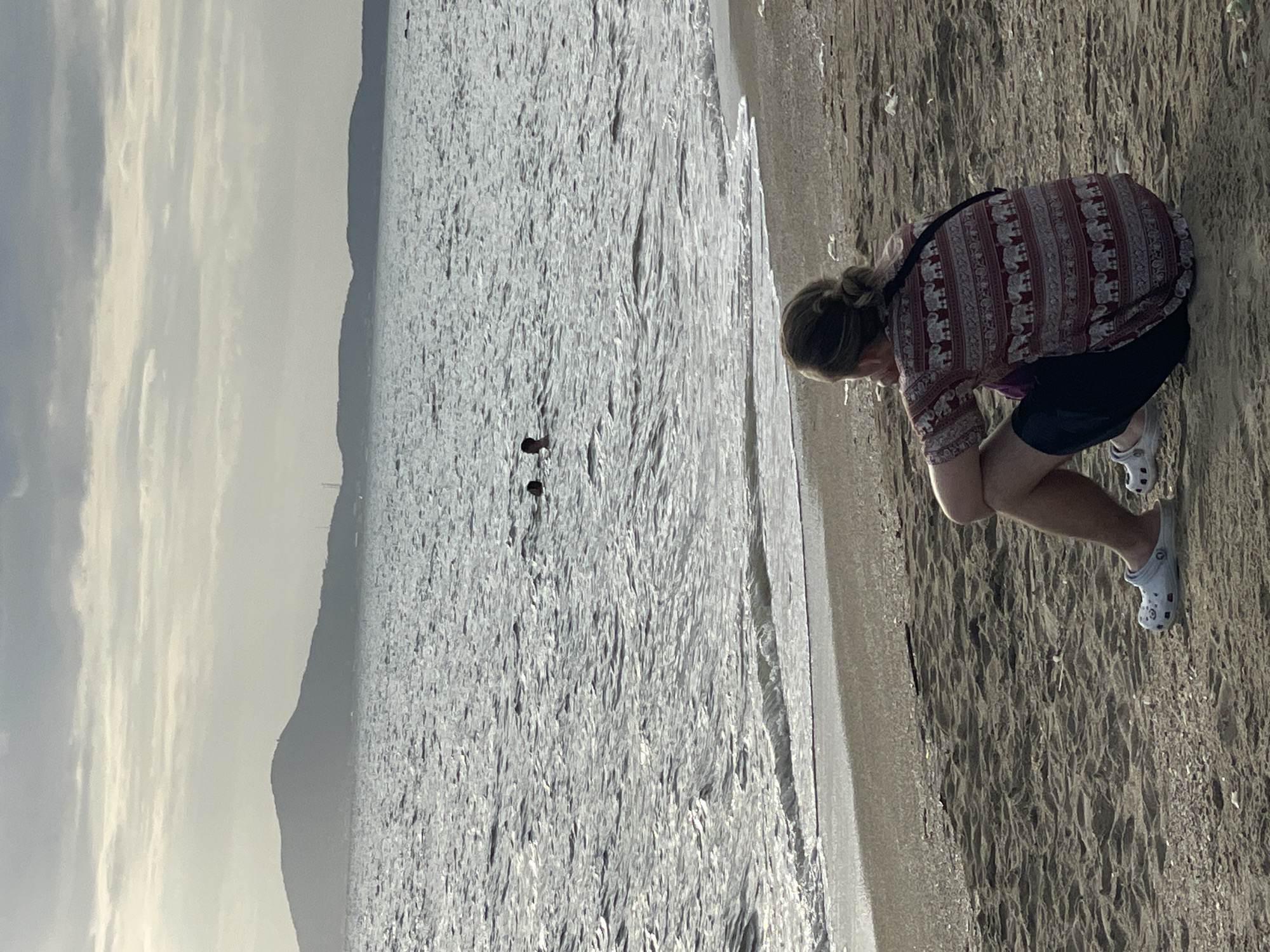 In Pattaya City, Thailand, a person with blond hair tied in a bun is seated on a sandy beach, facing away from the camera. They are wearing a patterned shirt with elephants and dark shorts, along with white clogs. The person has a black bag slung across their shoulder. In the distance, two people are swimming in the ocean. The sea is choppy, and a gentle wave breaks on the shore. In the background, a hazy mountain range with some structures is visible against a cloudy sky.