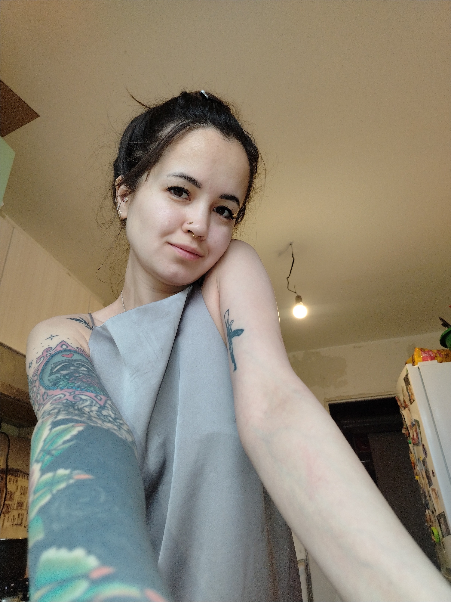 A young woman with dark hair pulled back in a bun and visible ear and nose piercings stands indoors. She has extensive tattoos on her left arm and a small tattoo on her right arm. She is holding a grey cloth. The background shows a kitchen area with cabinets, a refrigerator covered in magnets, and a hanging light bulb. The location is unspecified within the city of Noviy, Russia.