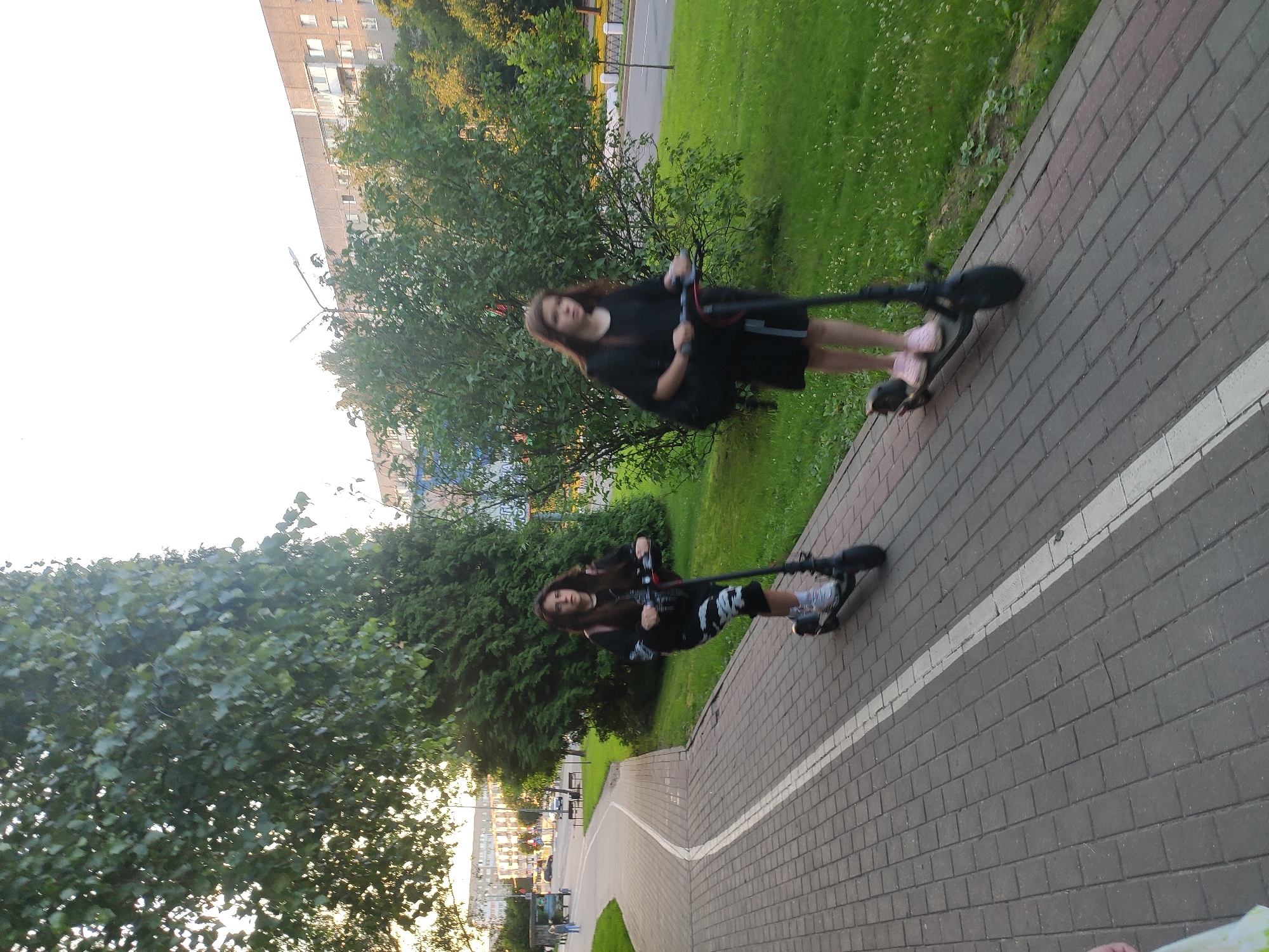Two young females are riding electric scooters on a paved walkway in Navapolatsk, Belarus. The walkway is bordered by a white line and has a grass area with trees and bushes on the right. In the background, there are apartment buildings and a street with cars and a lamppost. The sky is overcast. The first female, on the left, is wearing a black hoodie and patterned leggings. The second female, on the right, is wearing a black dress and pink sneakers. Both are looking forward as they ride.