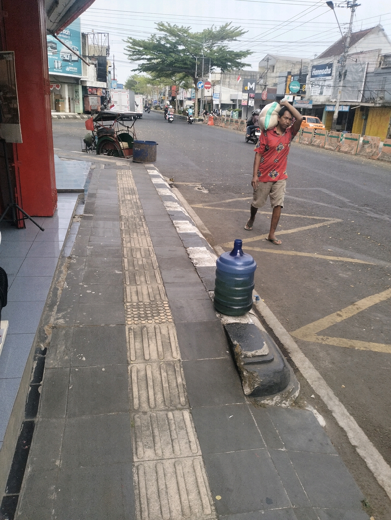 The image depicts a street scene in Purbalingga, Indonesia. In the foreground, a person is walking on a sidewalk, carrying a large sack over their shoulder. Beside the sidewalk, on the edge of the road, sits a large blue water dispenser jug. The street itself is paved and appears to be a busy thoroughfare with several vehicles visible in the background, including motorcycles and cars. Buildings line the street, displaying various signs and storefronts, suggesting a commercial or mixed-use area. The sky is overcast, indicating potentially dull weather. The overall impression is of a typical urban environment in Indonesia, with everyday activities taking place.
