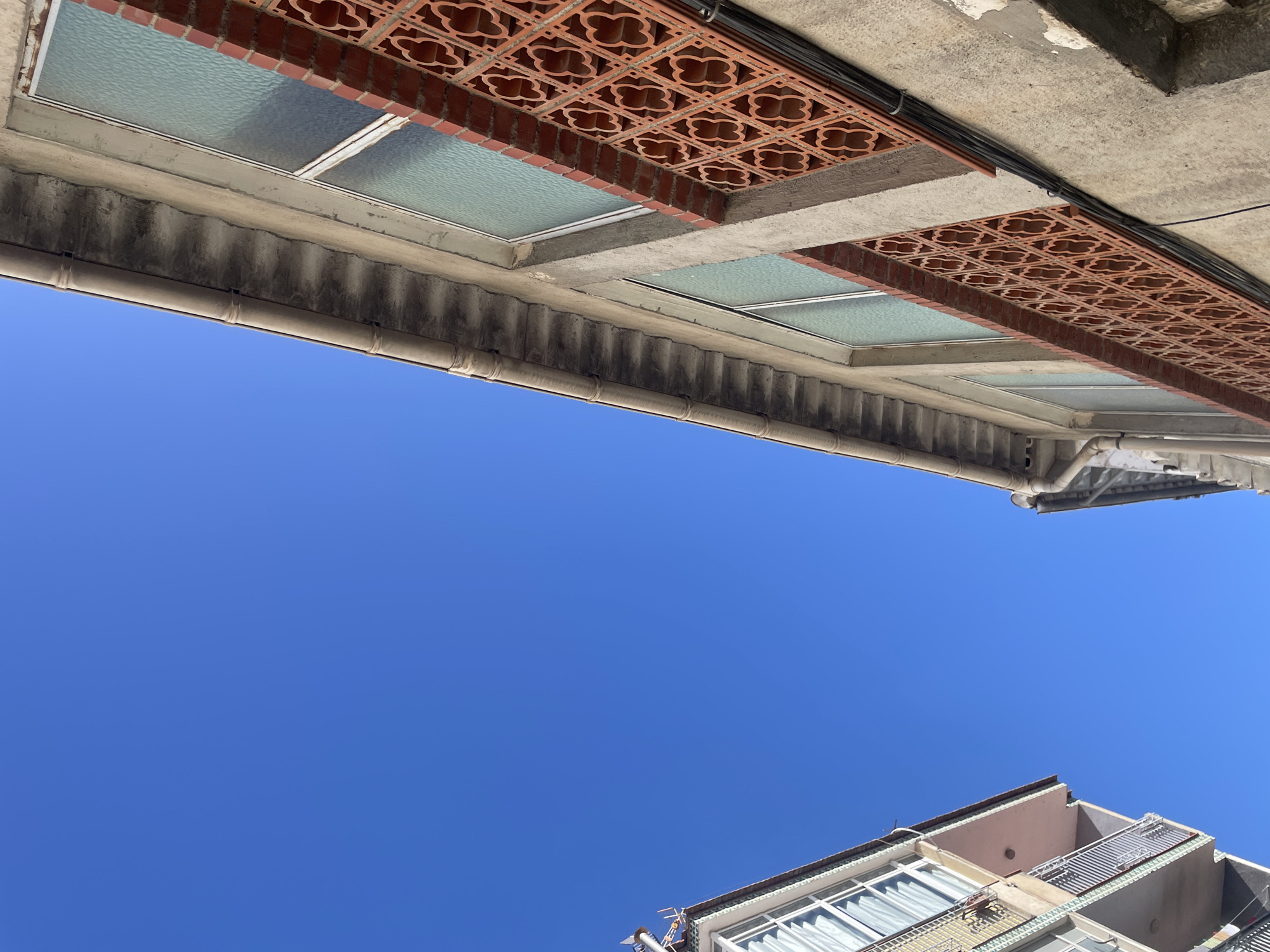 This is a low-angle, outdoor, daylight photograph taken in the city of Atarfe, Spain, looking up at two apartment buildings against a clear blue sky. The building on the right is closer and shows a concrete facade with a row of windows above a decorative terracotta lattice panel. The lattice has a repeating pattern of four interconnected, four-leaf clover shapes. Above the windows is a concrete gutter system. The building on the left is further away and is lighter in color, showing balconies with railings and windows. Electrical conduits are visible running along the side of the building on the right.