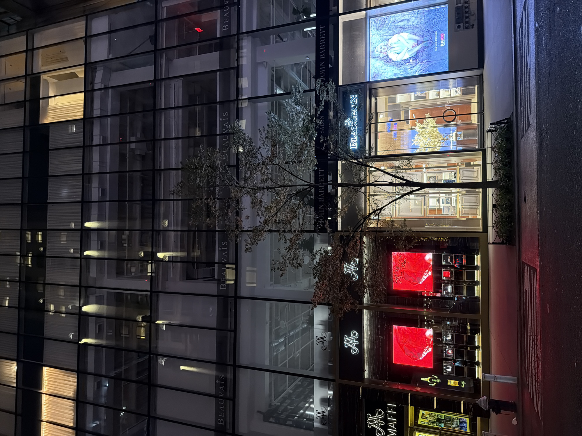 This image captures a nighttime street scene in New York City, likely on a commercial block. The focal point is a modern building with a glass facade, reflecting the city lights and interior illumination. Several storefronts are visible at street level, displaying illuminated signage and merchandise. Among the visible signs are "BEAUVAIS," "MAFFI," "JOHN BARRETT," and "BLANCPAIN." Two prominent red screens are displayed in one of the storefronts, and a large digital billboard featuring a person is visible to the right. A bare-branched tree stands in front of the building, and a fire hydrant and a crosswalk are present on the sidewalk in the foreground. The overall ambiance is one of urban sophistication and commerce at night.