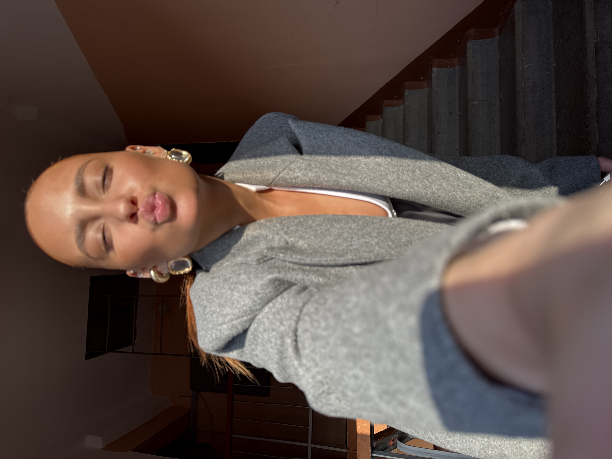An indoor, full-frame selfie captures a female subject in a stairwell. Her head is tilted slightly forward, with eyes closed and lips pursed in a kissing gesture. She exhibits a tanned complexion and light reddish-brown hair, illuminated by direct sunlight, which also casts bright highlights on her face and clothing. She wears large, gold-toned oval earrings with clear central stones, and multiple small ear piercings are visible on her left ear. Her attire includes an open grey textured blazer or coat over a white low-cut top. The subject's right arm, holding the camera, is blurred and partially visible in the lower foreground. The background consists of a stairwell featuring dark concrete steps and risers. The walls are painted a uniform reddish-brown hue. A dark metal railing is visible on the left side, paralleling the stairs. Sunlight creates strong highlights and defined shadows throughout the scene. The specific location within the city of Chaikovsky, Russia, cannot be determined from the visual information provided.