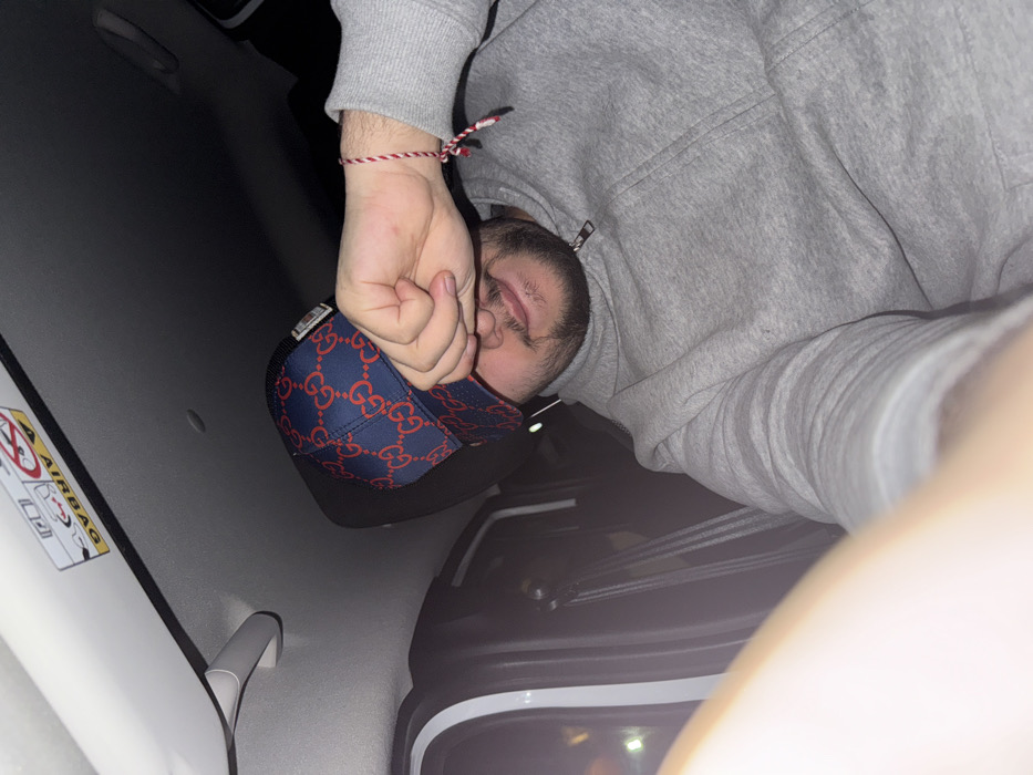 The image depicts a male individual seated in the passenger side of a vehicle, appearing to be at an unspecified location within Vellmar, Germany. The male subject is observed from a slightly low angle, positioned towards the right side of the frame, partially obscuring his face with his left hand. He has a dark beard and mustache, and his mouth is subtly curved in what appears to be a slight smile. He is wearing a grey zippered hooded sweatshirt and a dark blue baseball cap with a red "GG" (Gucci) pattern, worn backward. On his left wrist, a red and white braided string bracelet is visible.
The immediate environment is the interior of a car. The white ceiling panel of the vehicle is visible in the upper half of the image, featuring a grey grab handle on the passenger side. An "AIRBAG" warning sticker, displaying German text and international symbols indicating airbag and child seat hazards, is affixed to the sun visor. The vehicle's dark-colored interior upholstery is partially visible behind the subject. To the left side of the frame, part of a white car door frame and a dark window are visible, beyond which blurry, yellow-white external light sources suggest an outdoor, low-light, possibly nighttime setting. The subject's right arm and torso occupy the lower portion of the image.