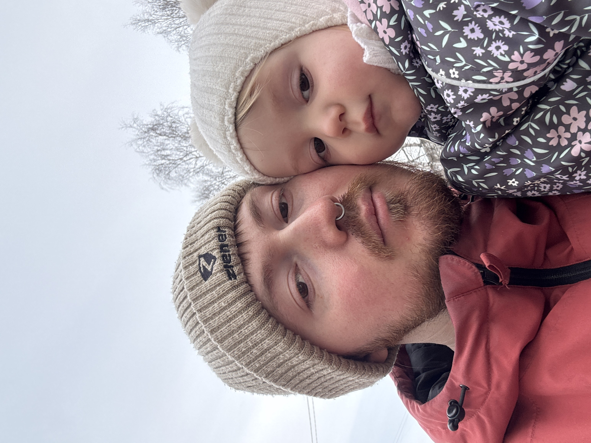 A low-angle shot shows a man and a toddler in what appears to be an outdoor setting. The man is wearing a light brown knit beanie with a dark logo on the left side and a nose ring in his left nostril. He has a reddish beard and is wearing a red jacket. The toddler is wearing a white beanie with a pom-pom and a floral-patterned jacket. The sky appears overcast. The location is Chaikovsky, Russia. Bare trees are visible in the upper left corner. The man and toddler are close together with the toddler appearing to be leaning on the man.