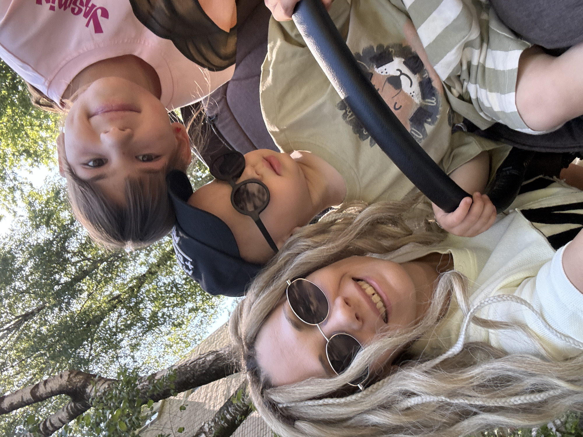 From a low angle, the image shows a selfie taken by a woman with blonde dreadlocks, wearing sunglasses, and smiling. Behind her and to the left is a girl with dark hair and a pink t-shirt. Next to the girl is a toddler in a black cap and sunglasses. Another child, wearing a beige t-shirt with a lion design, is in a stroller behind the toddler. The stroller has a black handle bar. Green trees and sky can be seen in the background. The location is identified as Rusanovo, Russia.