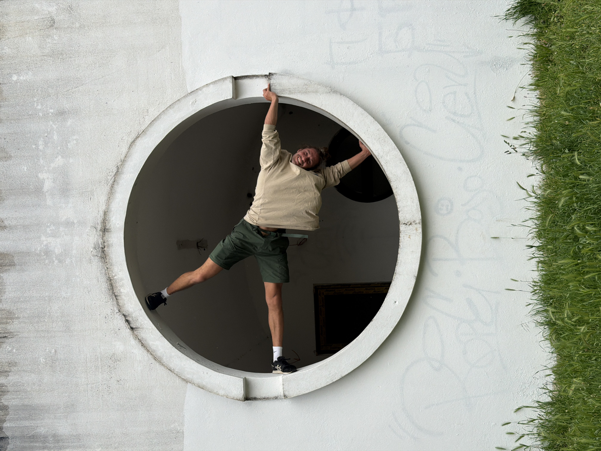 A man is hanging in a circular window in Petrovac centar, Montenegro. The man is wearing a beige long-sleeved shirt, green shorts, black shoes with white socks, and is smiling. He is grasping the top of the window with his right hand. His left arm is extended outwards, with the hand grasping the frame. His left leg is extended outwards, and his right leg is dangling down inside the window. The wall is white with some graffiti and some spots. To the right, there is a strip of green grass. Inside the window opening, a black frame is visible.