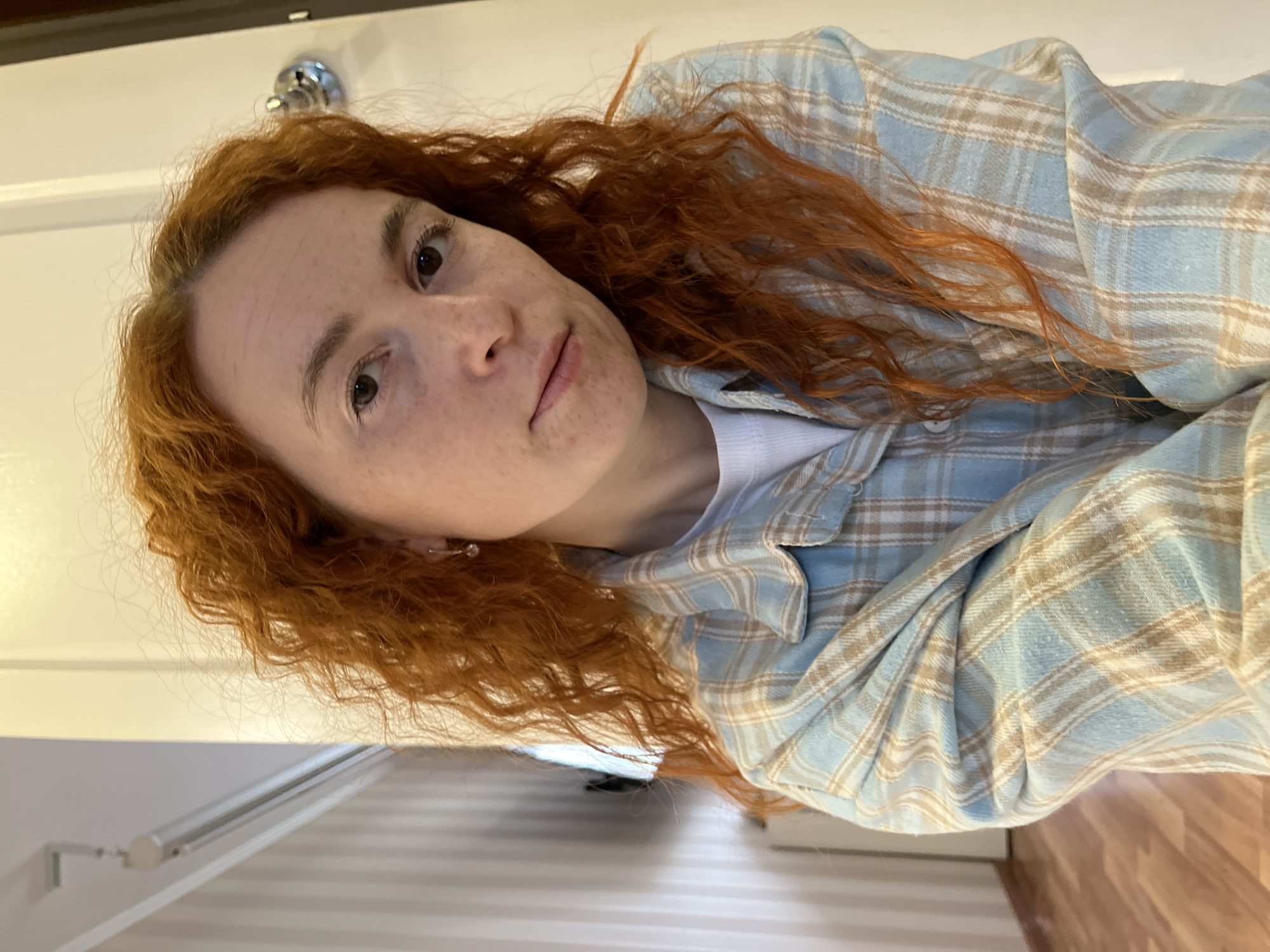 A woman with reddish-brown, curly hair is shown wearing a light blue and beige plaid shirt over a white t-shirt. She is looking at the camera with a neutral expression. The background includes a white door with a silver knob, a wall with a white striped pattern, and a wooden floor. No information to deduce that the location is in the city of Chaikovsky, Russia.