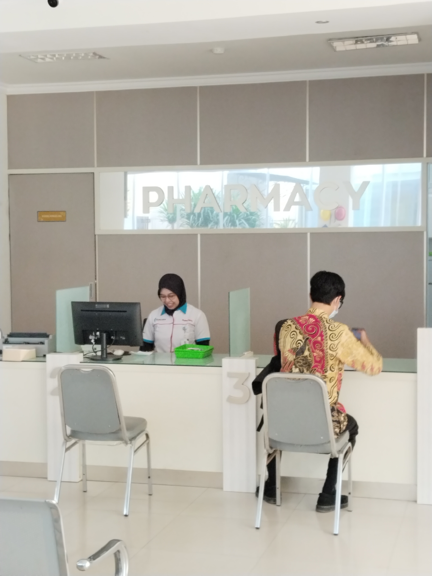 In a pharmacy in Kecamatan Pati, Indonesia, two individuals are visible. A female pharmacy worker, wearing a hijab and a white uniform with blue trim, smiles as she sits behind a counter with a computer monitor and a green tray. To her right, a person wearing a batik shirt and a mask sits facing away from the camera, holding a phone. The pharmacy features a large "PHARMACY" sign above the counter and multiple chairs for waiting customers. The environment appears clean and well-lit, with a tiled floor and white walls.