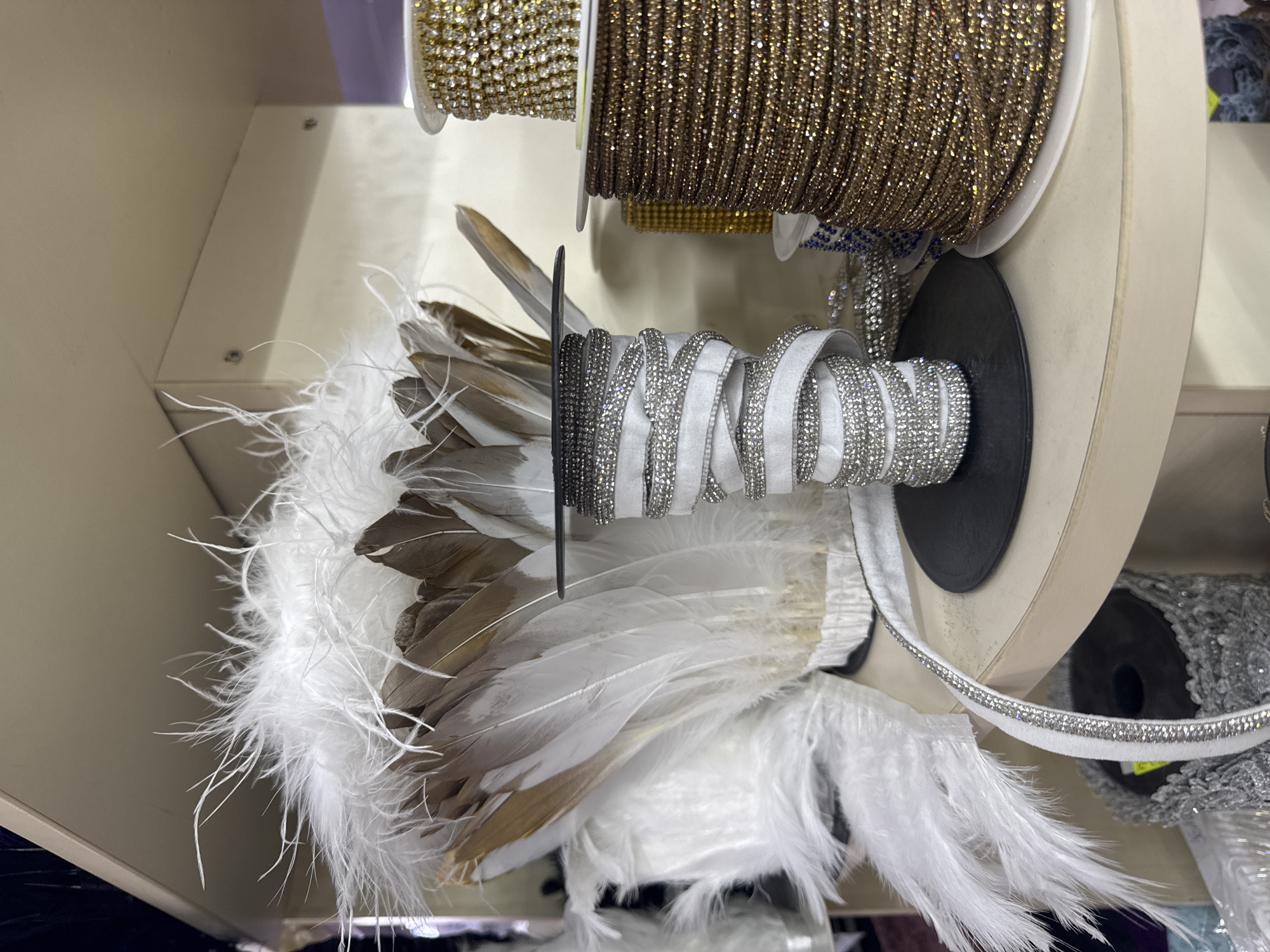 A close-up, angled shot shows a display of craft supplies, likely in a store, but the specific location in Sofia, Bulgaria, cannot be determined from the image alone. At the top, there are two spools of decorative trim - one with gold or yellow gemstones and another with brown gemstones. Below these are spools of white ribbon with silver gemstones wrapped around a spool holder, as well as additional spools of silver gemstones. Adjacent to these materials, a cluster of white and brown feathers is situated, possibly for use in decorations or costumes. The scene appears to be part of a retail or craft-oriented environment.