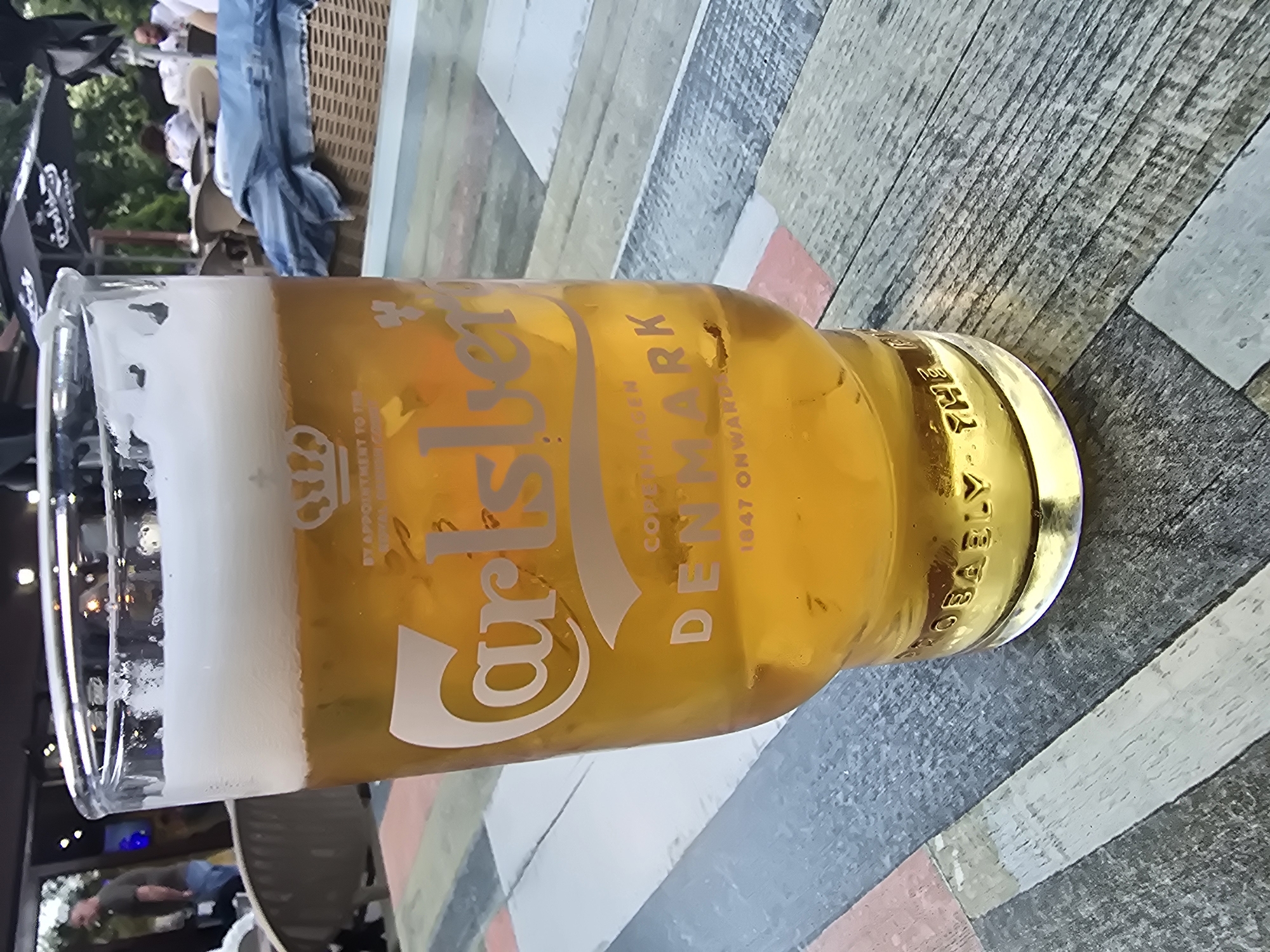 A close-up shot shows a glass of Carlsberg beer on a patterned wooden table. The glass is nearly full with a generous head of white foam. The beer has a light amber color. The Carlsberg logo and the text "COPENHAGEN DENMARK 1847 ONWARDS" are printed on the glass in silver. The wooden table has a modern, slightly weathered appearance with alternating grey, white, and brown planks. In the background, partially visible through a window or opening, are figures of people and possibly outdoor seating, suggesting a bar or restaurant setting.