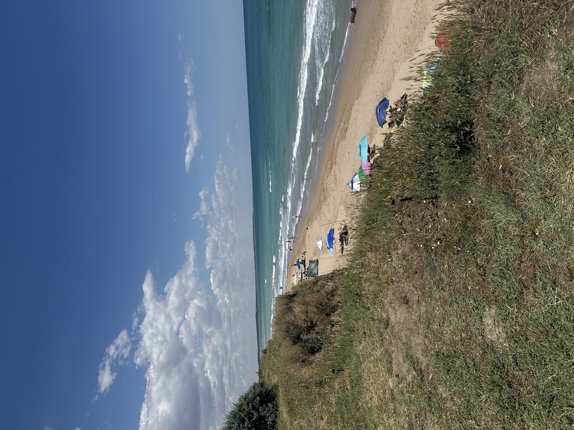 This image depicts a scenic view of a beach in Shabla, Bulgaria, on a sunny day with scattered clouds. The foreground features a grassy dune overlooking the sandy beach and the turquoise waters of the Black Sea. Several beach umbrellas are set up on the sand, providing shade for a few beachgoers. Some people are seen swimming or walking near the water's edge, while others are relaxing on the sand. The sea exhibits gentle waves, and the horizon stretches out to meet a clear blue sky. The vegetation on the dune is dry and sparse in some areas, indicating a warm, summer climate. The overall impression is one of a tranquil and inviting coastal environment.