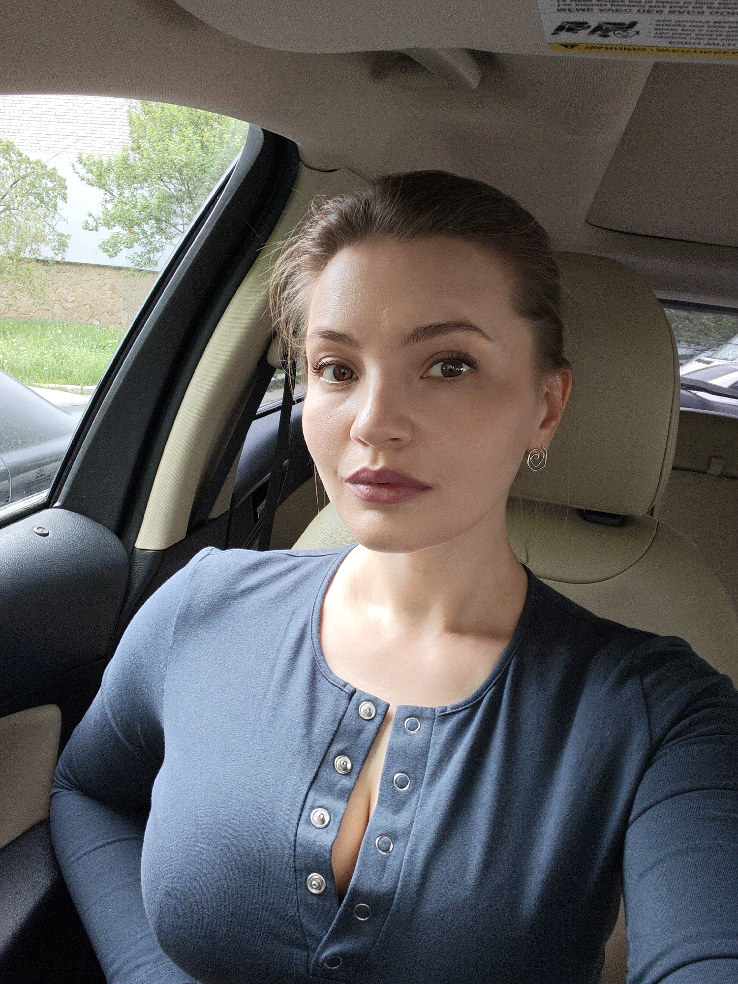 A woman is taking a selfie inside a car. She has light brown hair pulled back, brown eyes, and is wearing a blue button-down shirt. She is wearing small, coiled earrings. The car's interior is light colored. Outside the car window, trees and a building can be seen. The location is not discernible as being in Vinnytsia, Ukraine from the given image.