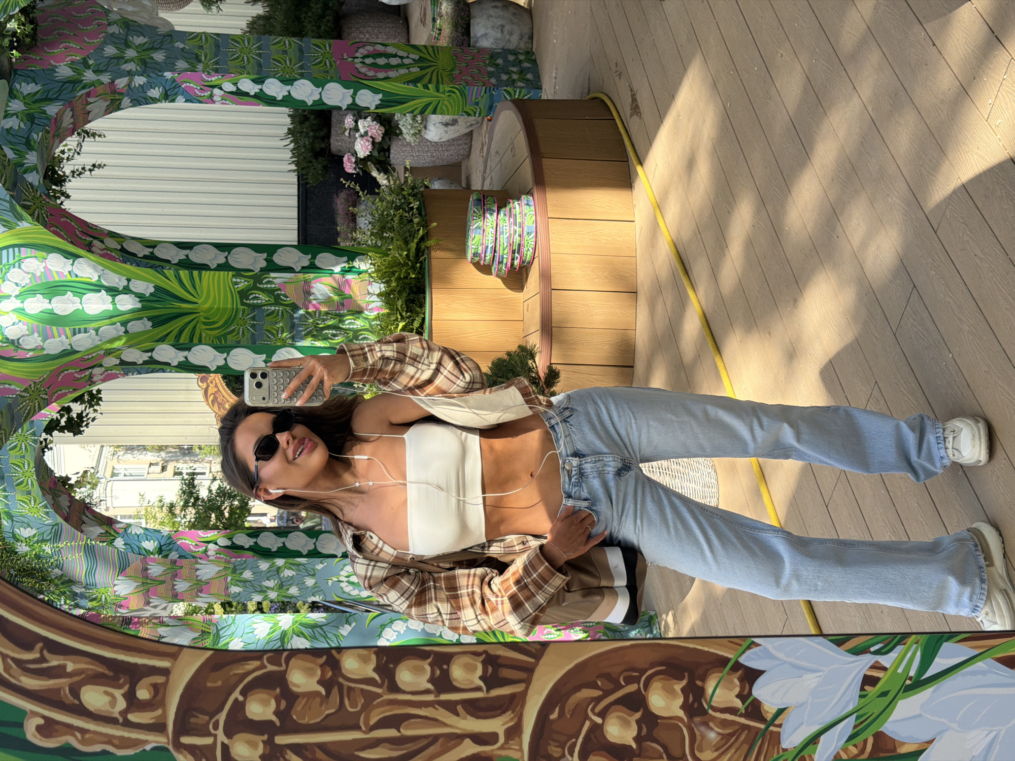 A young woman is depicted taking a mirror selfie in an outdoor setting. She is seen holding an iPhone in her hand, capturing her reflection in a large, ornately framed mirror. She is wearing a white tube top, light-wash jeans, and white sneakers, with a brown and white plaid shirt worn open over her top. Dark sunglasses obscure her eyes, and white wired earbuds are visible. A slight smile plays on her lips.
The mirror's frame is elaborately decorated with vibrant, intricate botanical motifs, predominantly featuring white lily of the valley flowers and other floral patterns in shades of green, pink, and blue, suggesting a lively, artistic installation. The ground beneath her is a light-colored wooden deck, marked by strong sunlight and shadows, indicating it is daytime and sunny. A yellow hose is visible lying on the deck.
In the background, visible both directly and reflected in the mirror, are various elements including wooden cylindrical structures that appear to be planters or benches, lush green plants, and clusters of pink flowers. A light-colored corrugated metal wall is also reflected in the upper part of the mirror. The overall scene suggests a leisurely moment in an aesthetically pleasing outdoor space, possibly a garden, patio, or an outdoor art exhibition in Moscow, Russia, where such public installations are common. There is no visible text in the image.