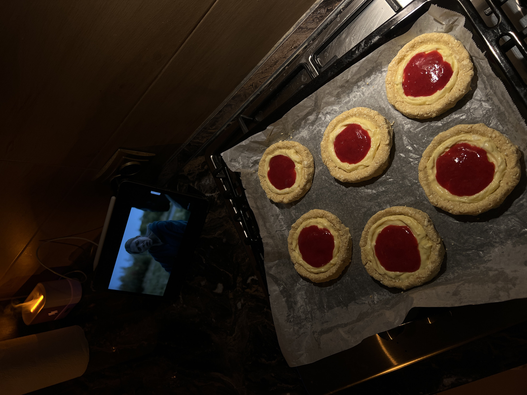 In what appears to be a kitchen in Ternopil, Ukraine, a tablet device displays a still image of a person with their mouth open in distress, set against a blurry outdoor background. The tablet rests on a dark, speckled countertop. Nearby, an oil diffuser emits a warm, orange light, resembling a flame.
In the foreground, a baking sheet lined with parchment paper holds six circular pastries. Each pastry has a golden-brown crust, a creamy yellow filling, and a dollop of red jam in the center. The pastries are arranged in two rows on the baking sheet, which is positioned on a stainless-steel stovetop. The ambient lighting is dim, with the primary illumination coming from the oil diffuser and the tablet screen.