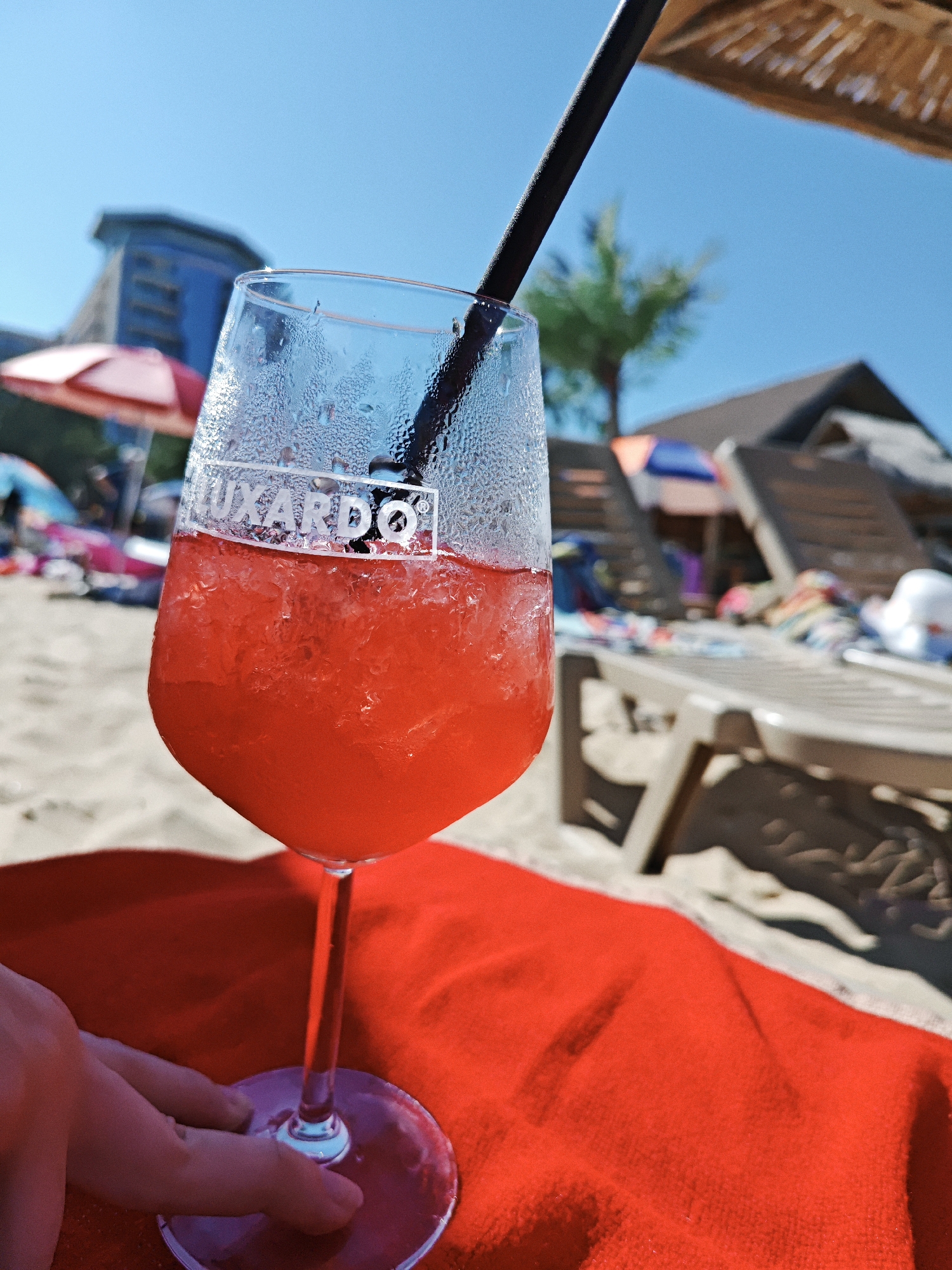 A close-up, low-angle shot focuses on a chilled red beverage in a clear wine glass filled with ice, topped with condensation, and featuring a black straw. The glass is branded with "LUXARDO" in white lettering. The drink rests on a bright red towel on a sandy beach. In the background, out of focus, are beach chairs, umbrellas, a palm tree, and a building, suggesting a resort or coastal setting. A person's hand is visible on the left, near the base of the glass. The sky is clear and blue.
