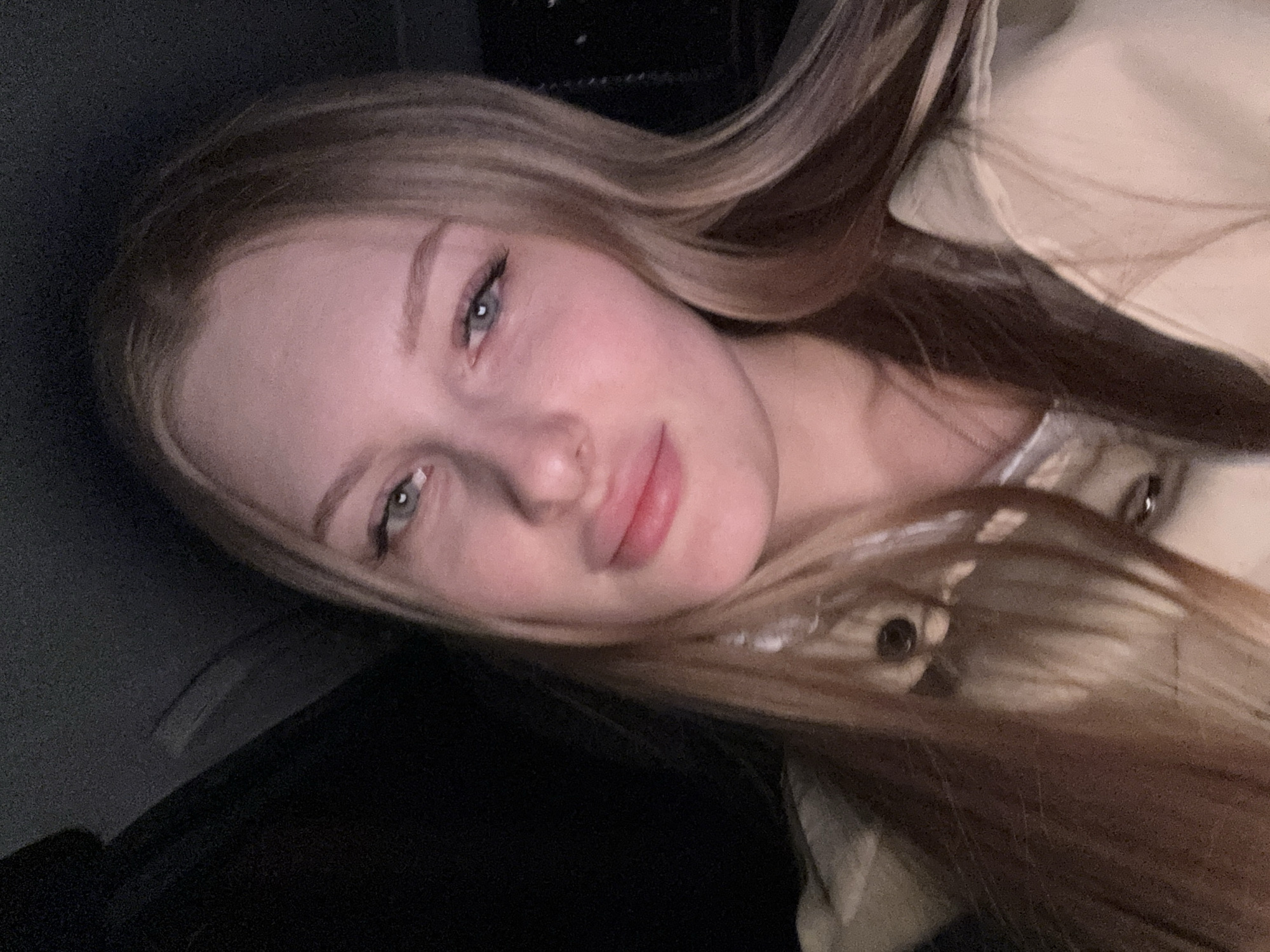 A young, fair-skinned woman with long blonde hair is the primary subject of the image. Her face is angled towards the camera, featuring light eyebrows, blue-grey eyes with dark eyeliner, a slightly pursed mouth, and a fair complexion. She appears to be wearing a light-colored jacket. The background is predominantly dark, with some indistinct shapes and textures. The photo's composition is a close-up, focusing on the woman's face. There is no indication of the location being Perm, Russia.