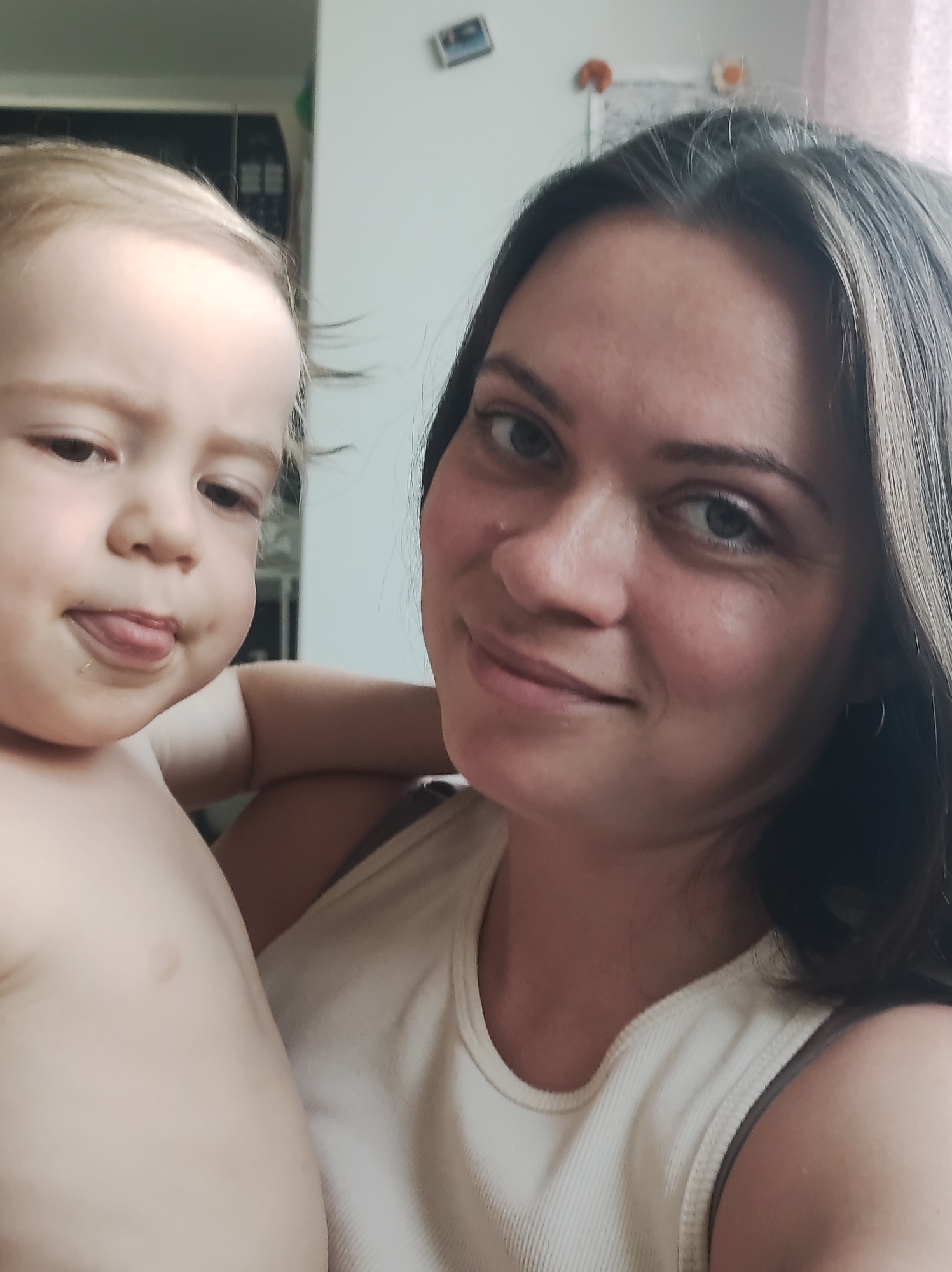 The image shows a light-skinned woman with dark hair holding a light-skinned toddler. The toddler is sticking out their tongue. The woman is wearing a light-colored tank top. A kitchen with appliances is visible in the background. The location is unknown, and the image does not provide any evidence of being in Chaikovsky, Russia.
