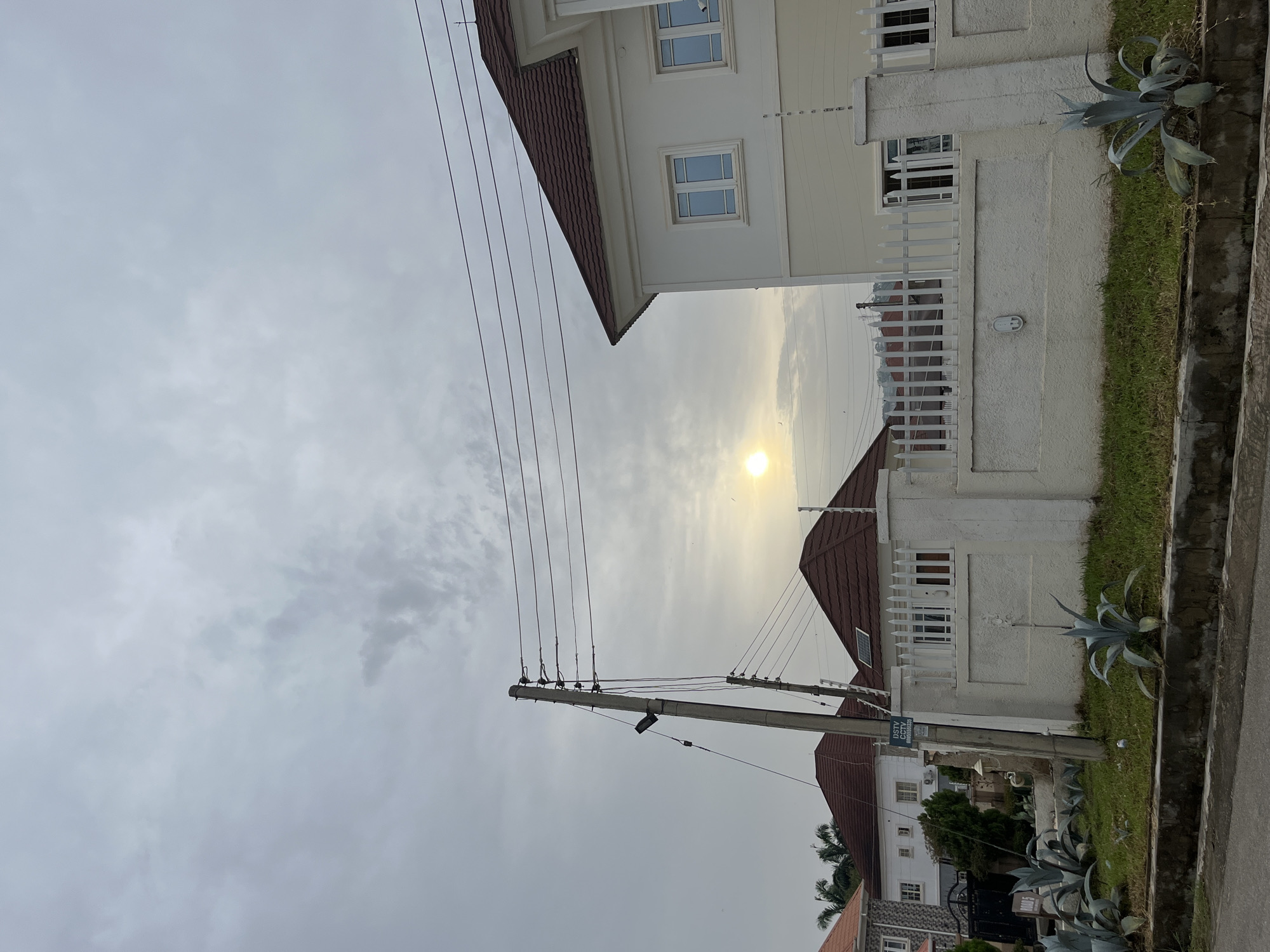 The image depicts a residential street scene in Abuja, Nigeria. A row of houses with white facades and brown tiled roofs lines the street. The sky is overcast, with the sun partially obscured by clouds. A concrete utility pole with numerous electrical wires is visible. Succulent plants are planted near the edge of the street. A sign with "DSTV" and phone numbers is attached to the utility pole.