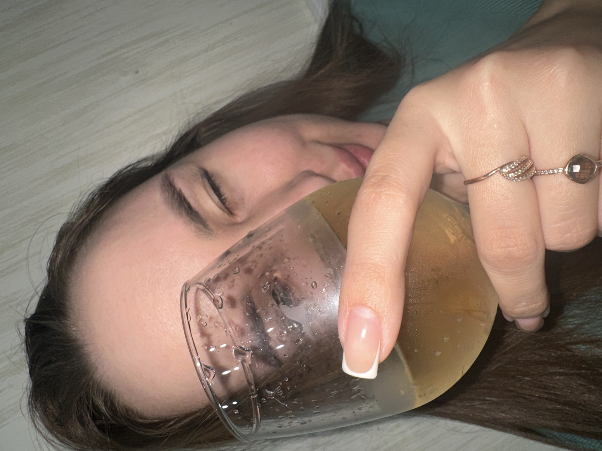 A woman is lying down while holding a glass of amber liquid. The woman has brown hair, closed eyes, and fair skin. She has two rings on her right ring finger. The glass has condensation on the outside. Part of the woman's hair is visible on a green surface. The background includes a beige wall. The location is likely indoors but the city of Chaikovsky, Russia, cannot be inferred from the image.