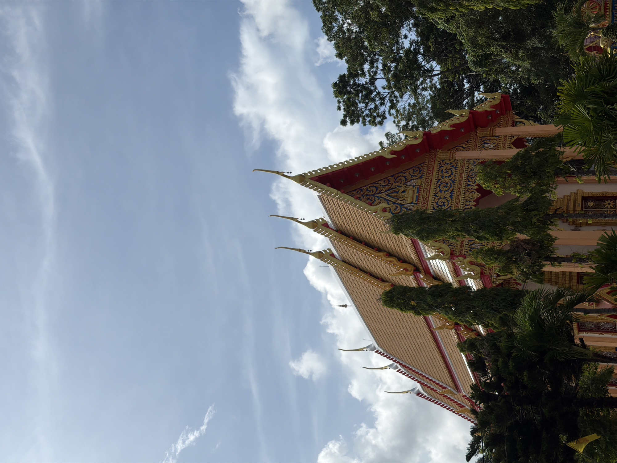 A Buddhist temple with intricate gold and red ornamentation on its roof is partially obscured by lush green trees. The sky above is a vibrant blue with scattered white clouds. The temple's architectural style, with its multi-tiered, pointed roofs and detailed decorative elements, is characteristic of temples found in Thep Krasatti, Thailand. The image captures a serene outdoor scene, with no people visible. The environment appears to be tropical, suggested by the dense foliage.